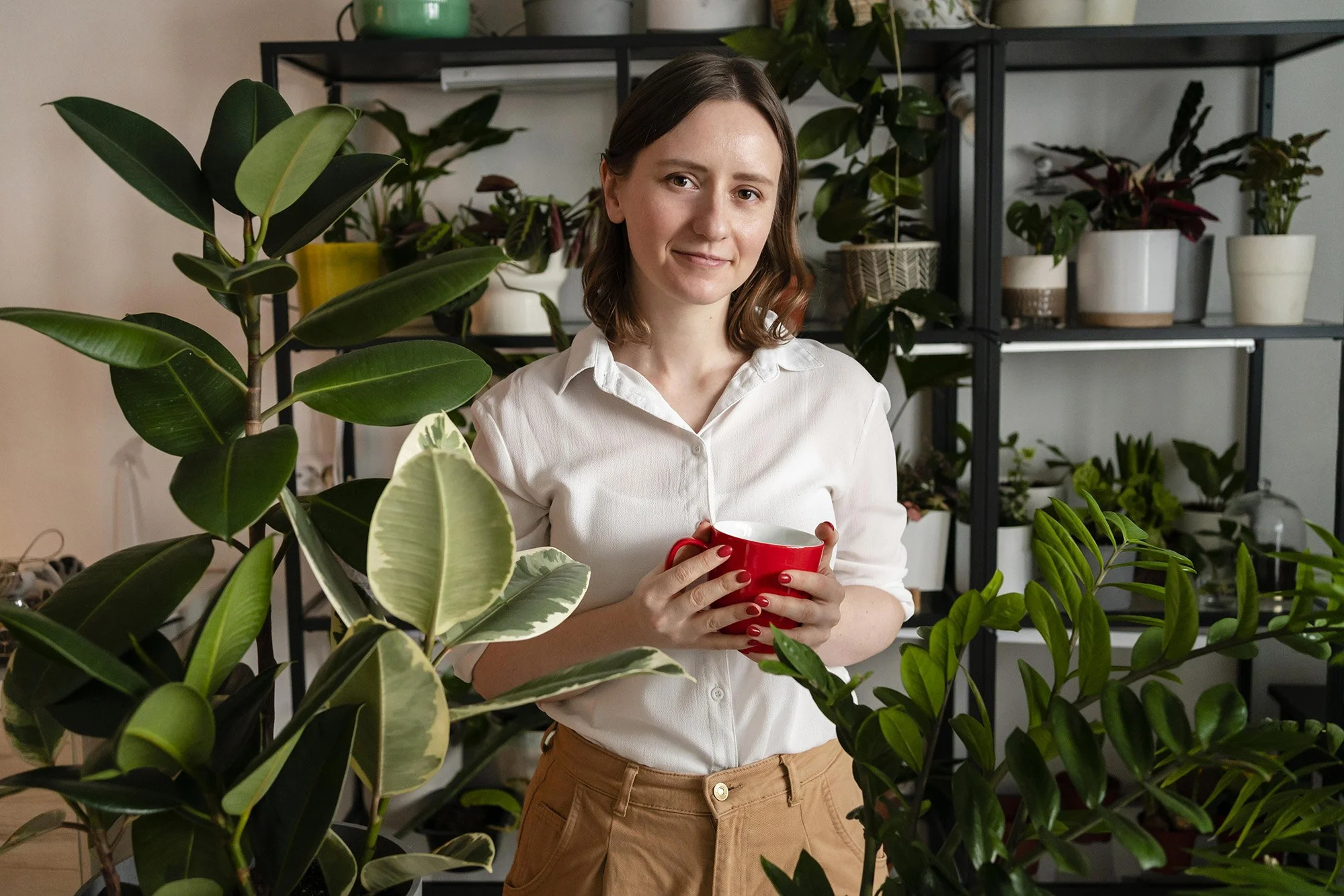 Una mujer joven con cabello castaño, vestida con blusa blanca y pantalones beige, sosteniendo una taza roja, rodeada de plantas en macetas y en un estante negro.
