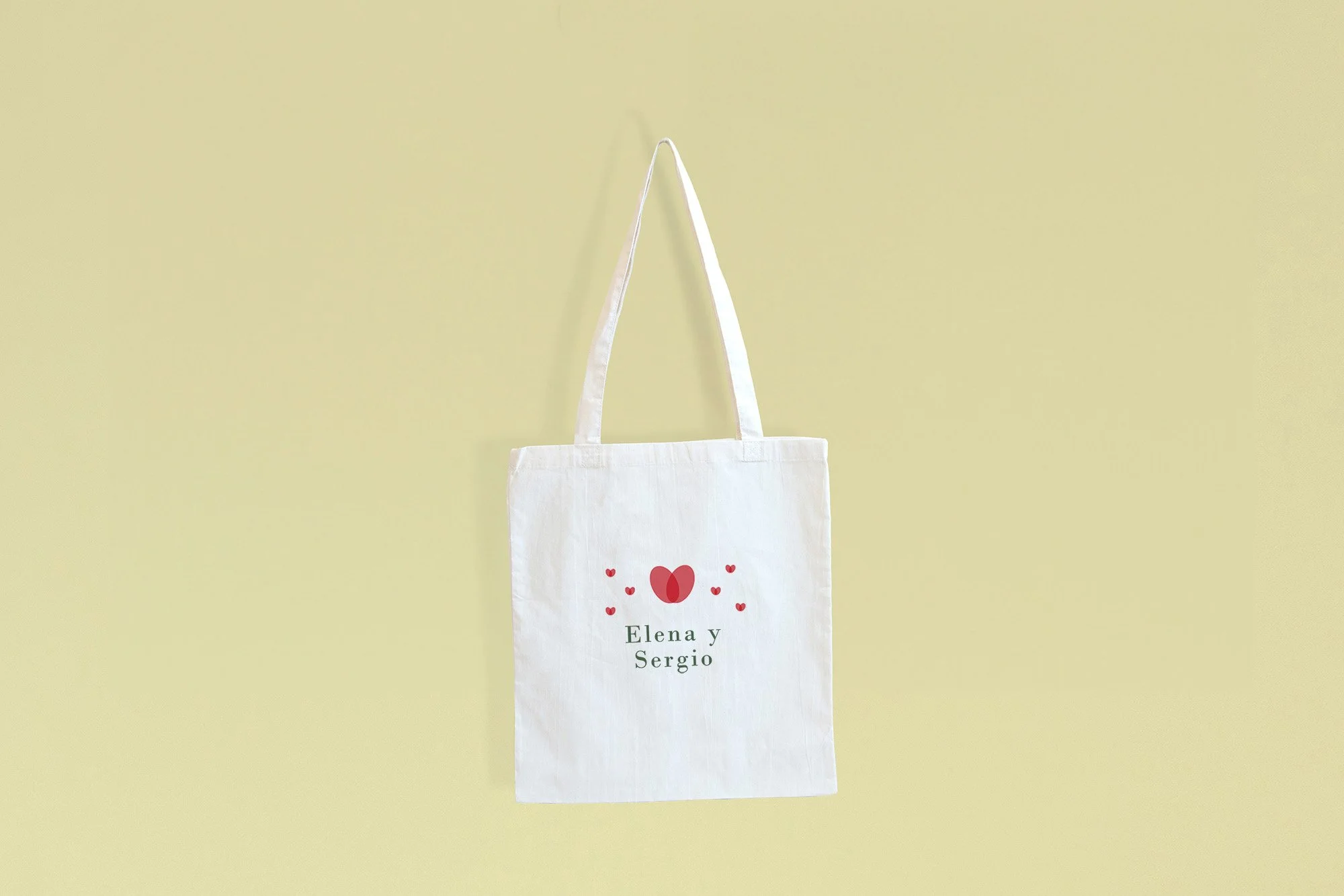 Bolsa de tela blanca con corazones rojos y nombres "Elena y Sergio" en letras negras, sobre fondo verde