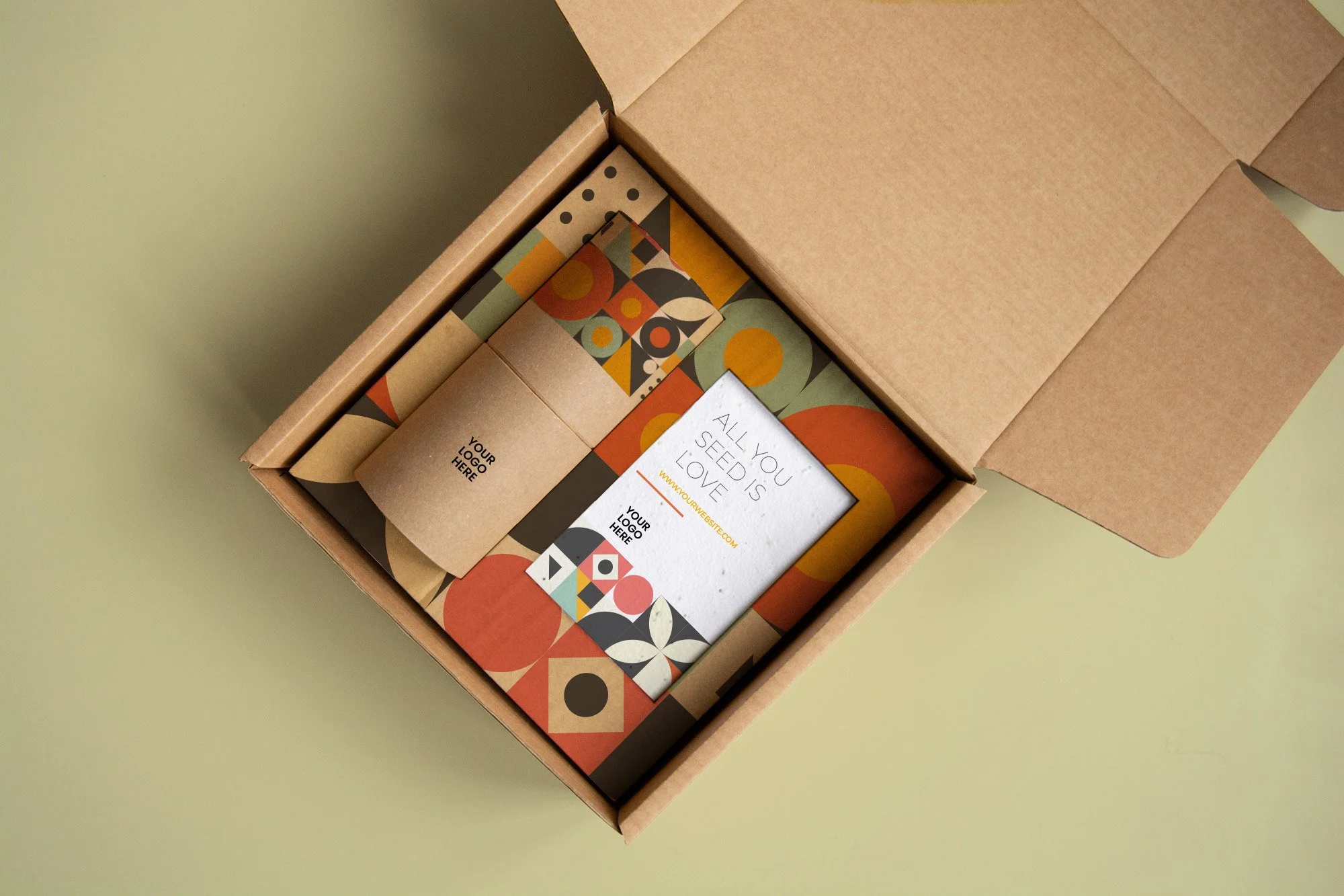 Caja de cartón abierta con materiales promocionales con diseños geométricos en colores naranja, negro, blanco y verde. Incluye un folleto con texto en inglés que dice 'All you seed is love' y espacio para logo y sitio web.