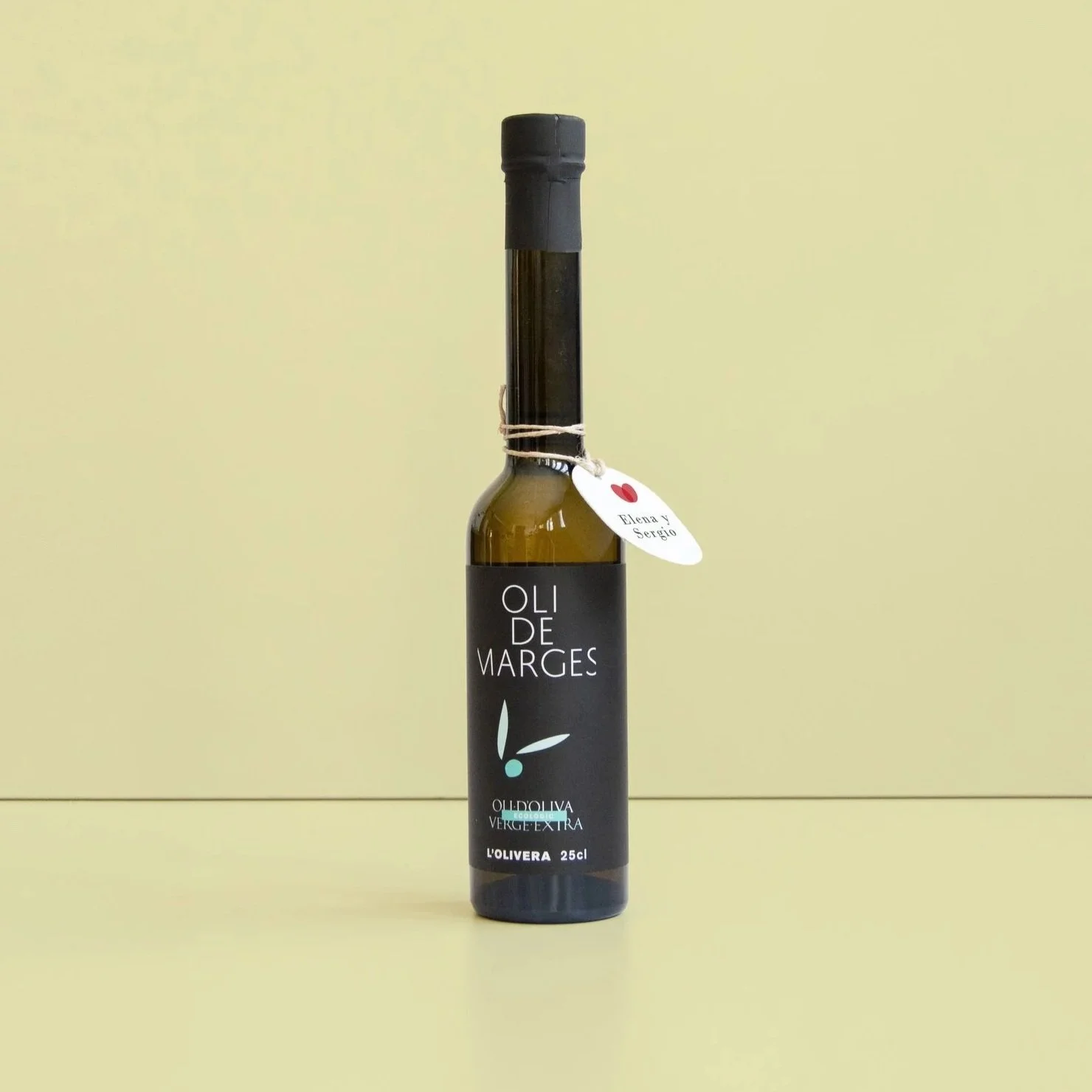 Botella de aceite de oliva con etiqueta negra, marca 'Oli de Marges', y un pequeño colgante con nombres y corazón.