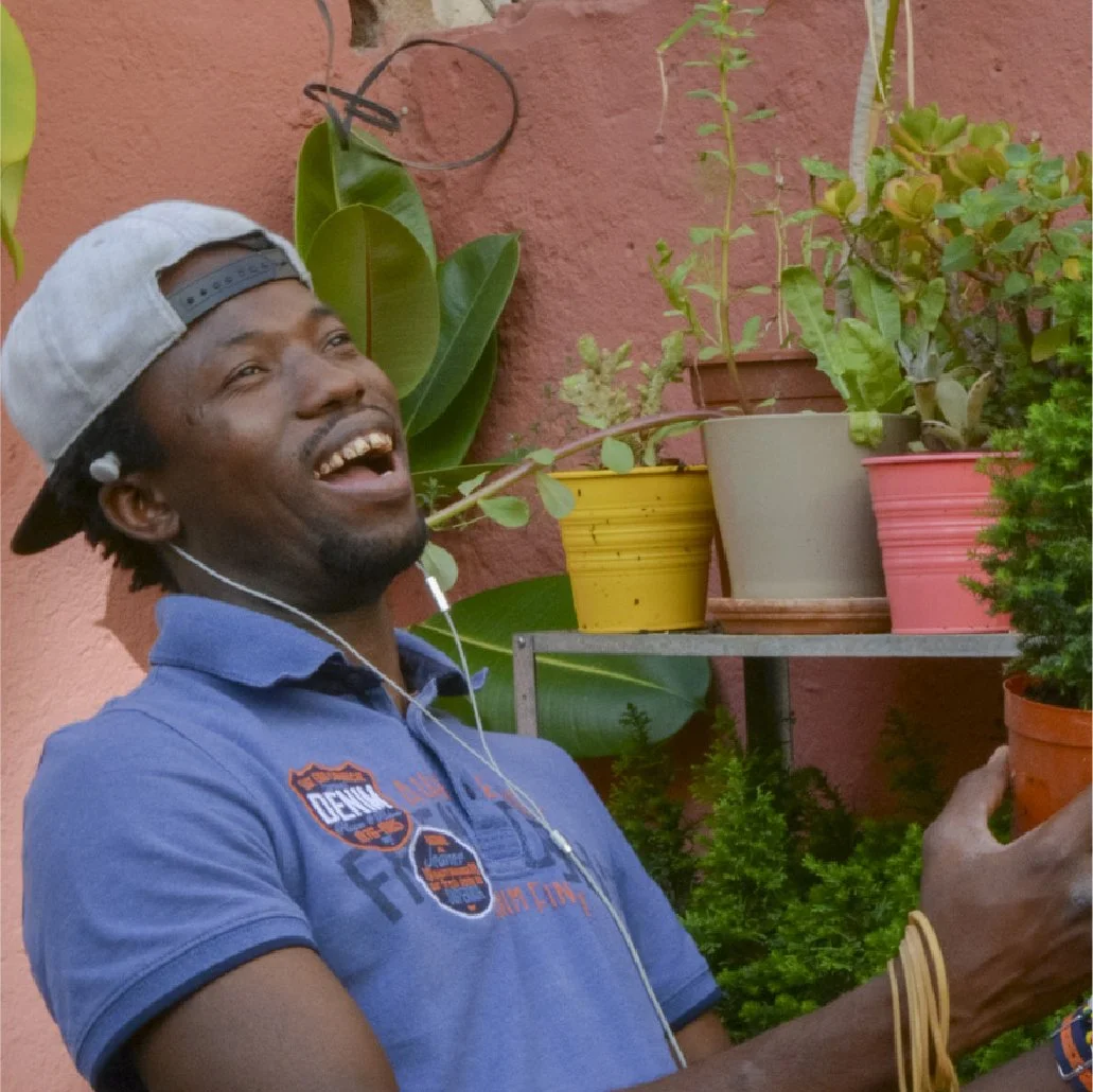 Un hombre sonriente con gorra mirando un macetero con plantas en un fondo de pared color terracota.