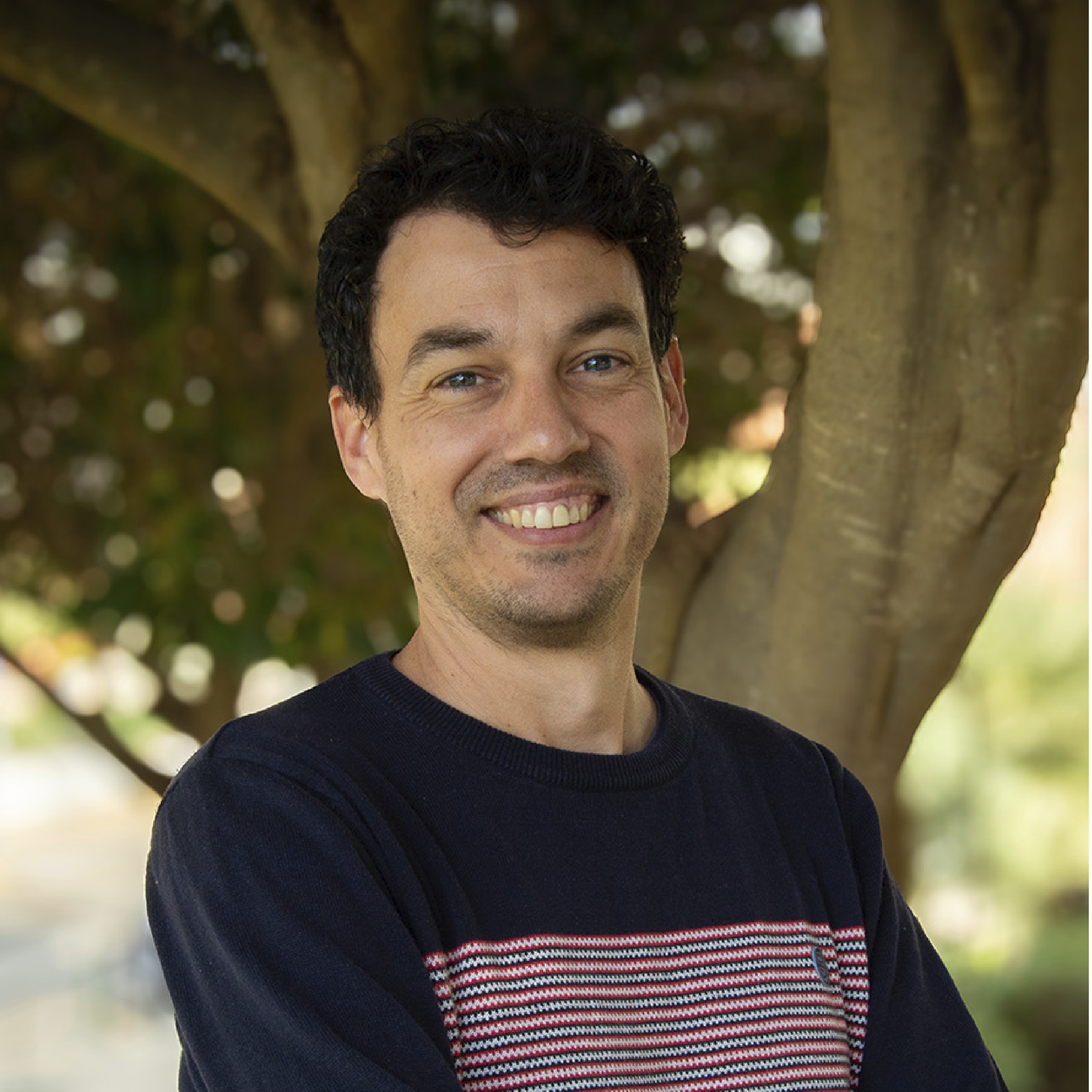 Retrato de un hombre joven con cabello rizado oscuro y ojos claros, sonriendo, con árboles y luz natural en el fondo.