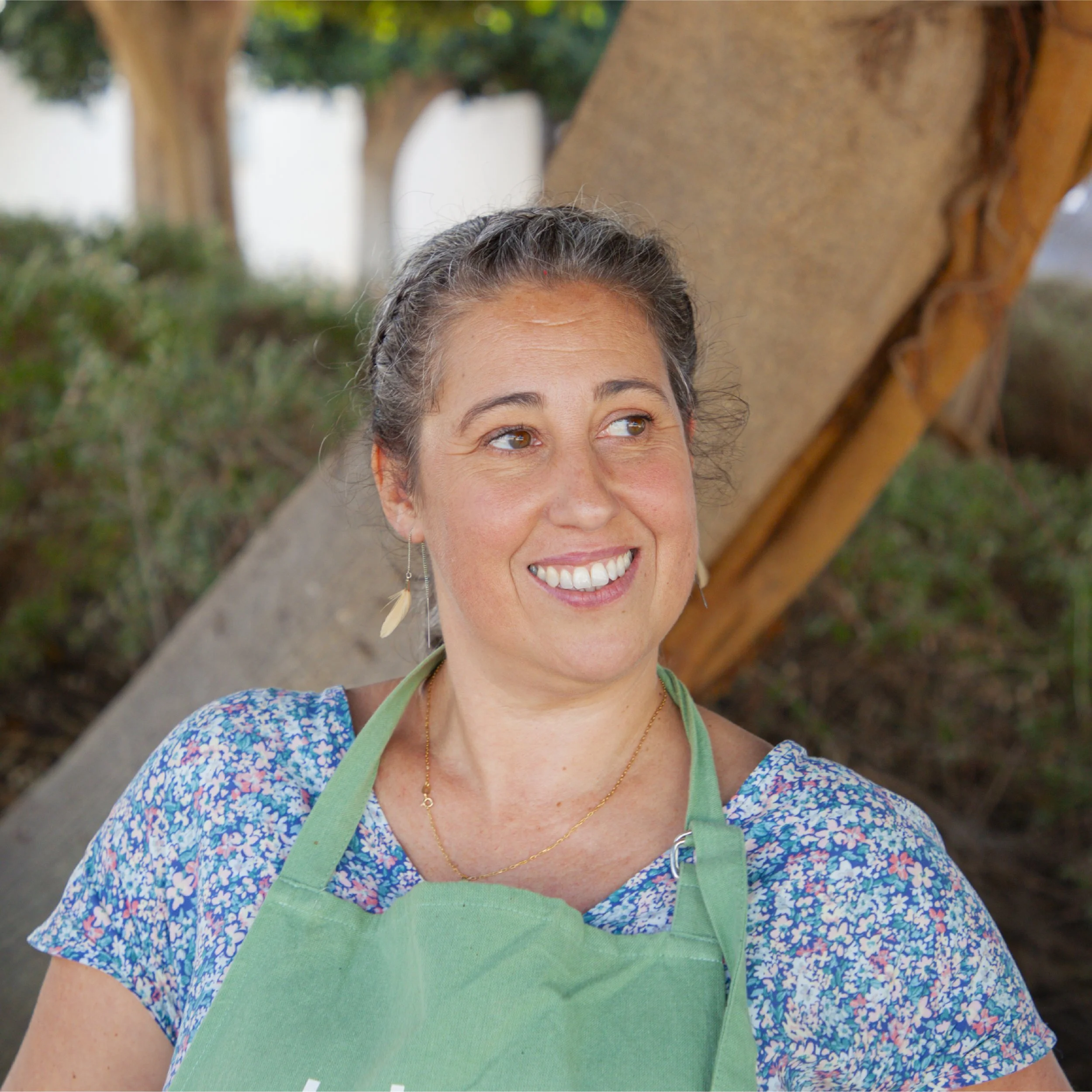 Mujer con cabello castaño claro con mechas grises y sonriendo, lleva camiseta azul con estampado floral y delantal verde, de fondo en un entorno al aire libre con árboles.