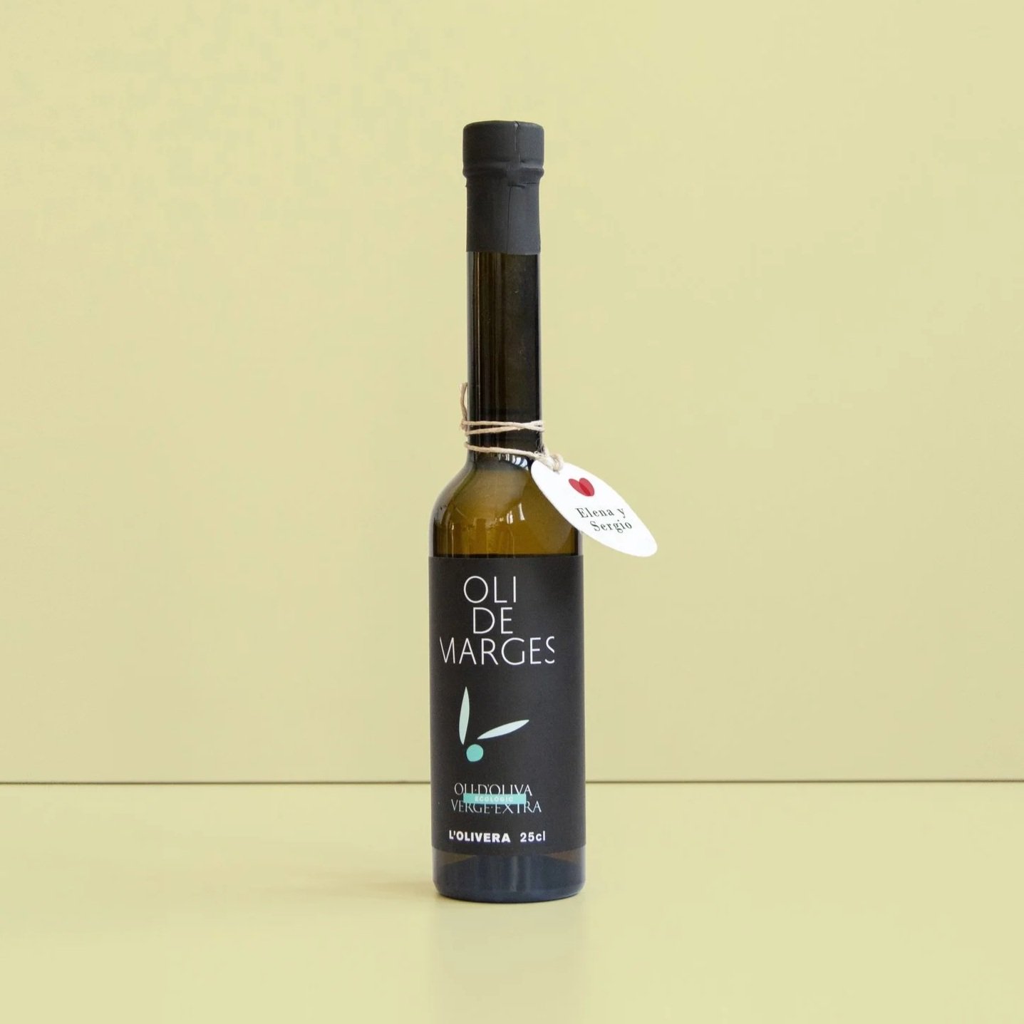 Botella de aceite de oliva con etiqueta negra que dice 'Oli de Marges' y un colgante con un corazón y los nombres Elena y Sergio.