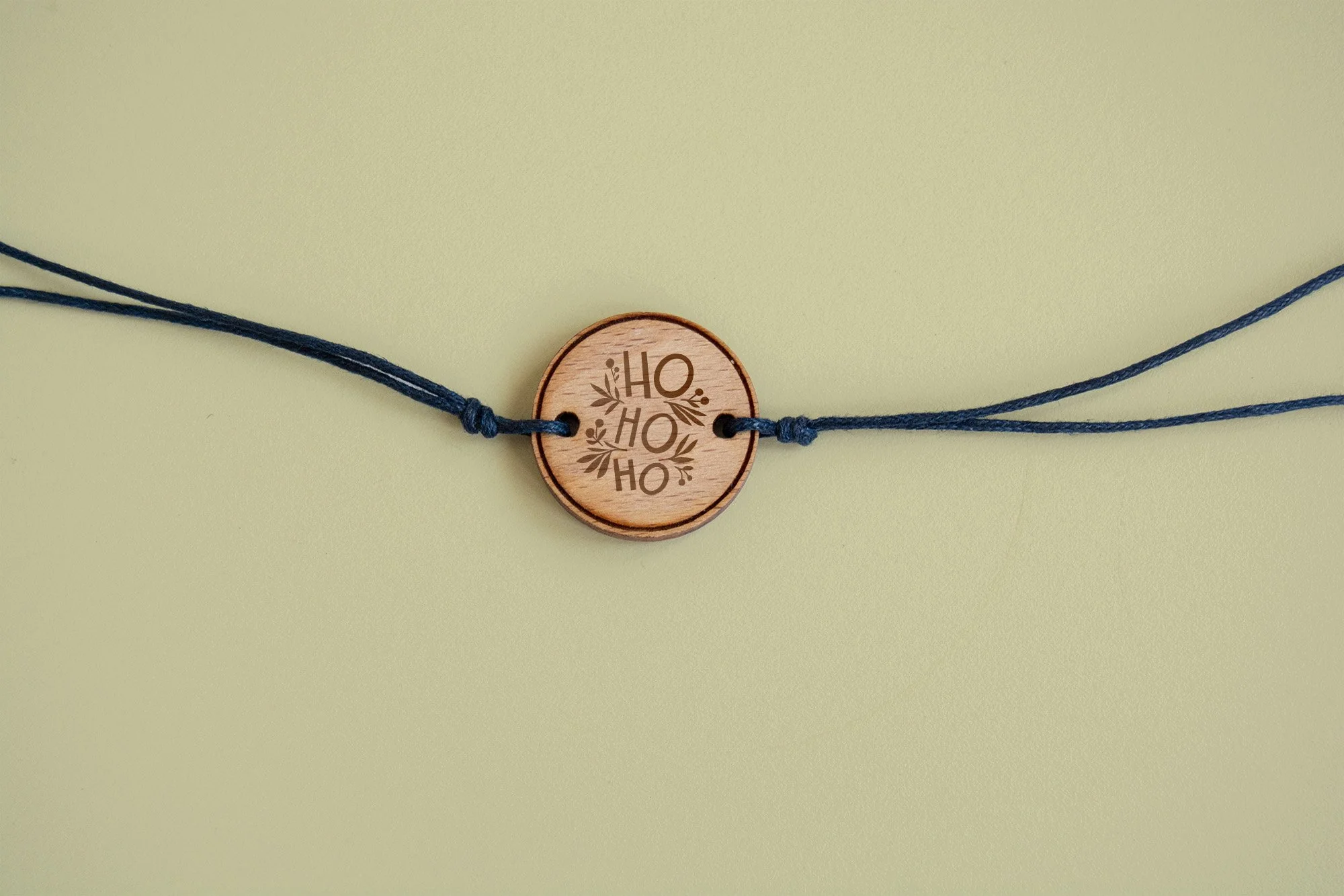 Collar hecho de cuerda azul con una pieza circular de madera que tiene grabado un mensaje que dice 'HO HO HO' rodeado de ramas de acebo.