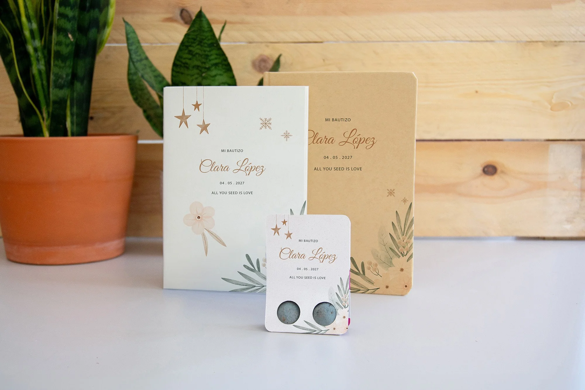 Tarjetas de bautizo con plantas y detalles decorativos, una en tamaño grande y otras más pequeñas, con fondo de pared de madera y una maceta con planta verde a la izquierda.