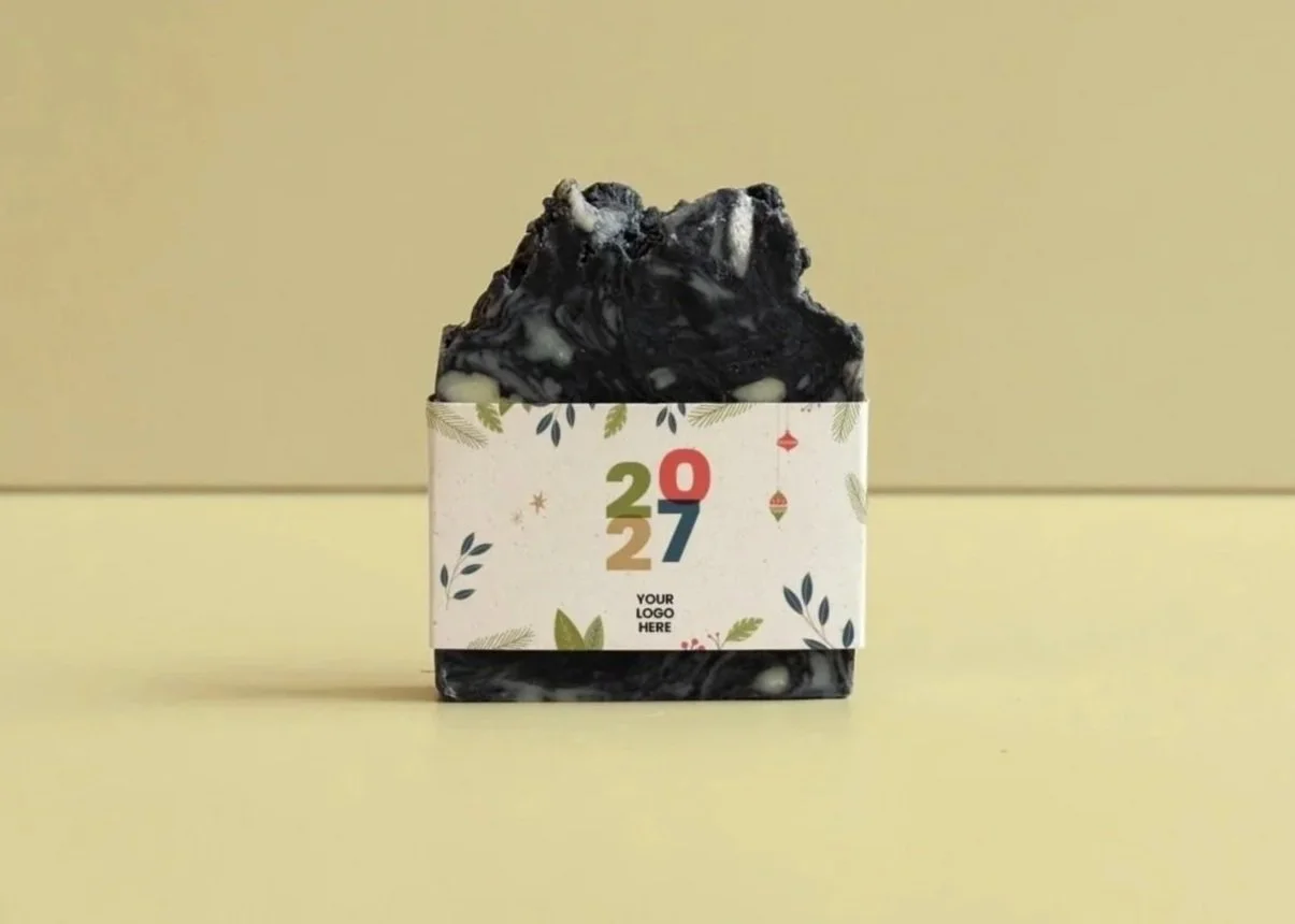 Jabón de color negro con crema blanca, envuelto con una cinta decorativa que tiene un diseño festivo y el texto '2027'.