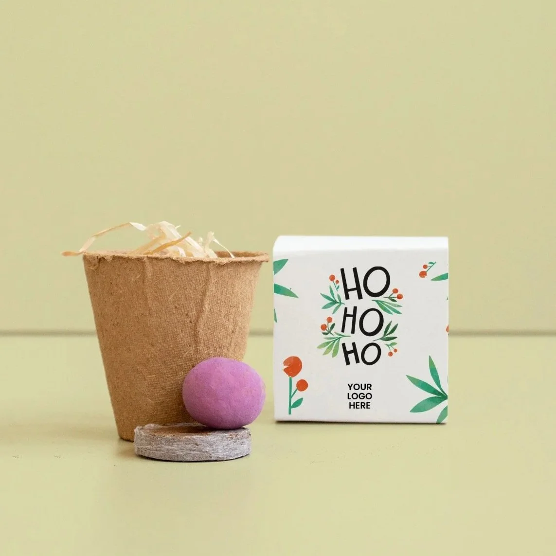 Una pequeña maceta de papel con relleno de rafia natural, una bola morada de semillas y un paquete con diseño navideño que dice 'Ho Ho Ho' y espacio para un logotipo.