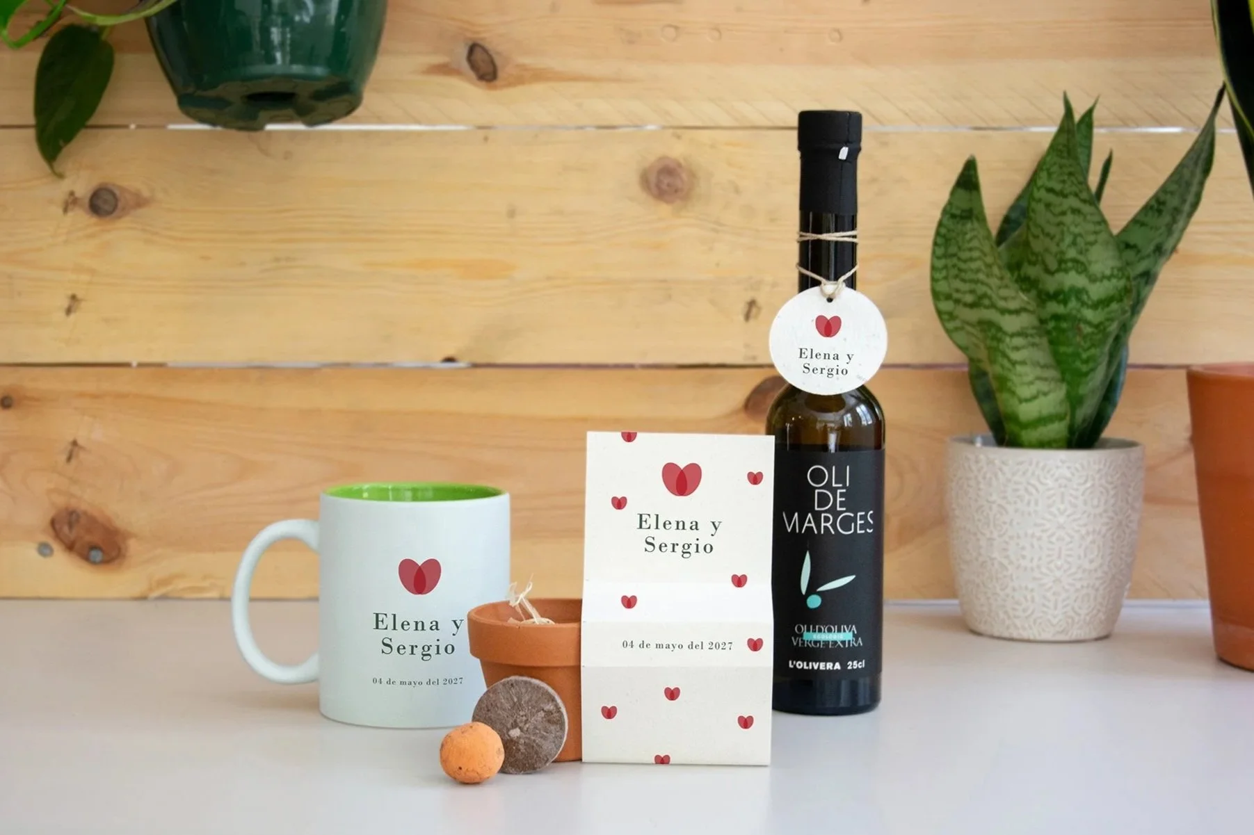 Una taza blanca con interior verde, una pequeña maceta de barro, una tarjeta con nombres y fecha, una botella de aceite de oliva, y dos plantas en macetas de cerámica, todo colocado sobre una mesa contra una pared de madera. Con diseño de bodas.