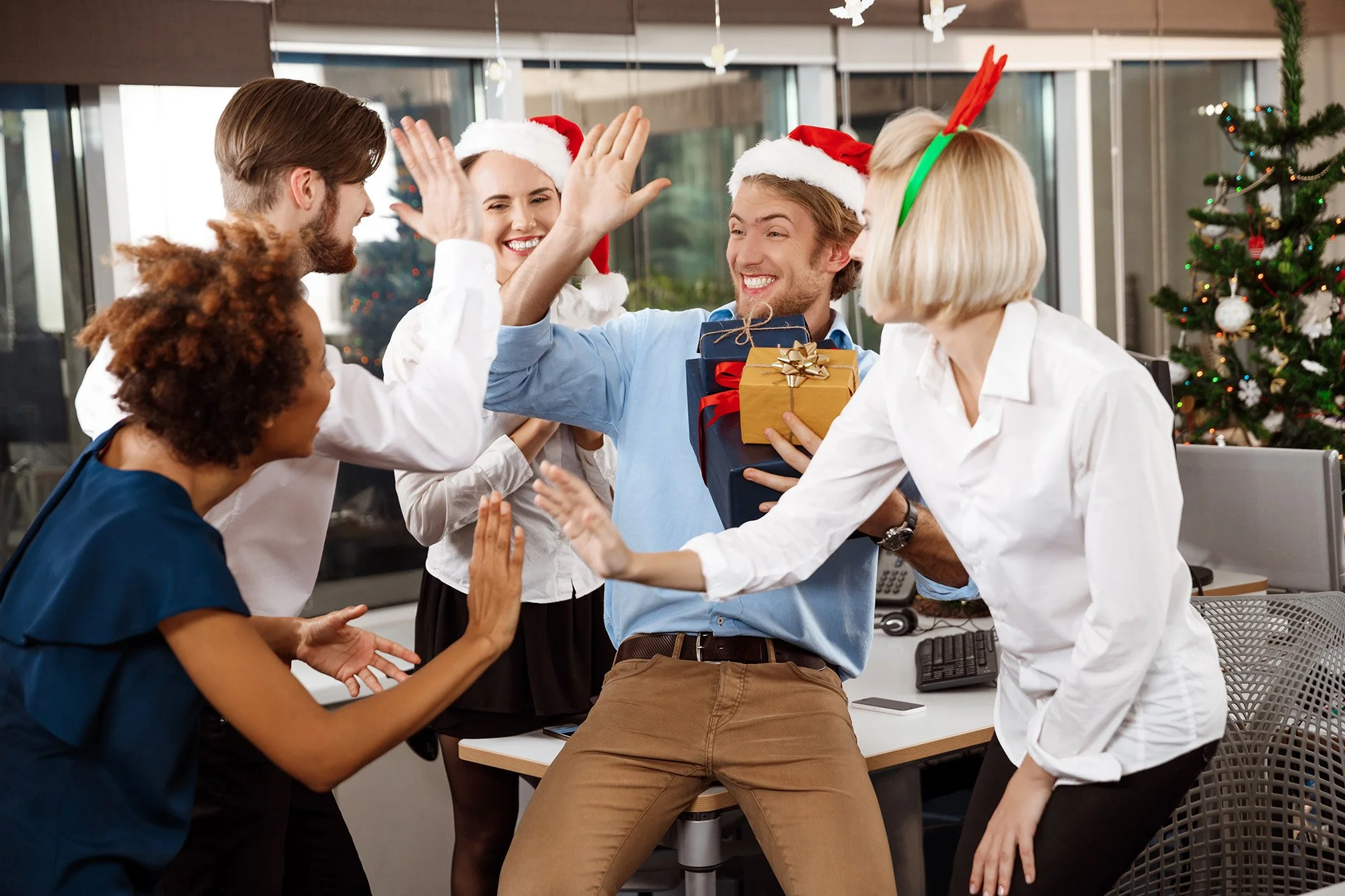 Grupo de personas en celebración navideña en una oficina, sonriendo y dándose palmadas, algunos llevan gorros de Santa y uno tiene una diadema de reno. En el fondo hay un árbol de Navidad decorado.