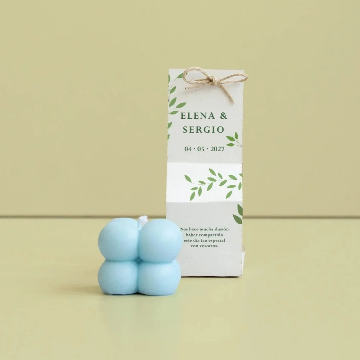Caja de regalo blanca con decoraciones verdes y texto, junto a cinco velas celestes en forma de burbuja, en un fondo verde.
