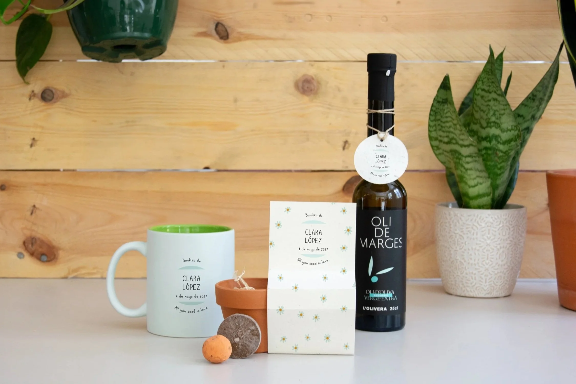 Una botella de aceite de oliva con una etiqueta que dice 'Oli de Marge', una taza blanca con la misma etiqueta, una tarjeta que menciona un bautizo de Clara López el 4 de mayo de 2027, varias plantas en macetas de cerámica y una pequeña bola de piedra, todo sobre una superficie blanca frente a un fondo de pared de madera.