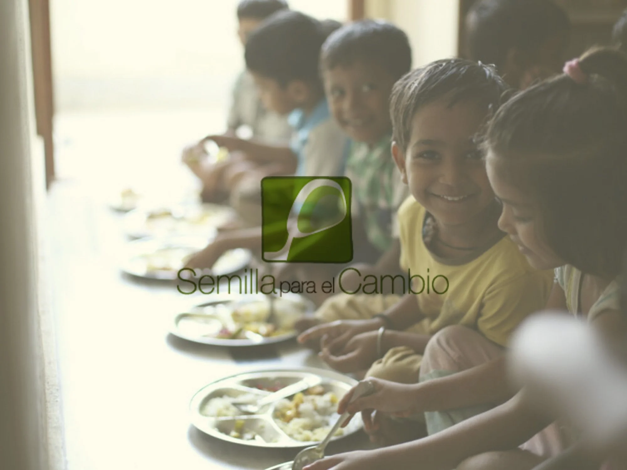 Niños comiendo en una mesa, algunos con platos de comida frente a ellos, en un ambiente escolar, con una sonrisa en el rostro.