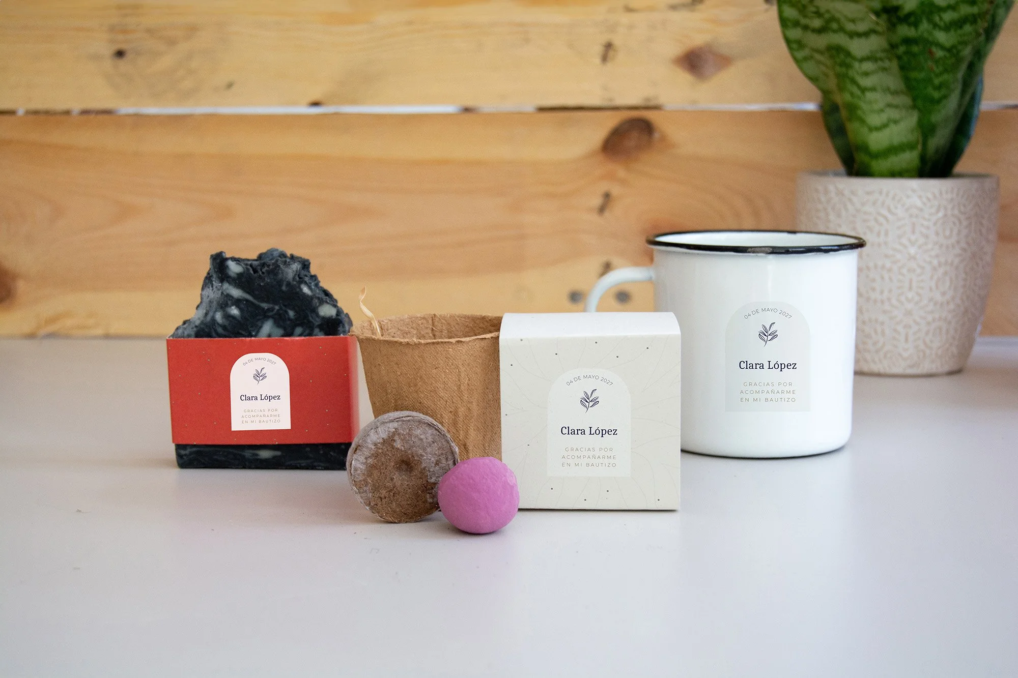 Conjunto de artículos personalizados, incluyendo una taza blanca con detalles en negro, unas tarjetas con nombres y agradecimientos, una piedra decorativa, un pequeño pom pom rosa, y un vaso de cerámica decorado, todo colocado sobre una superficie blanca contra un fondo de madera, con una maceta con planta en la esquina derecha.
