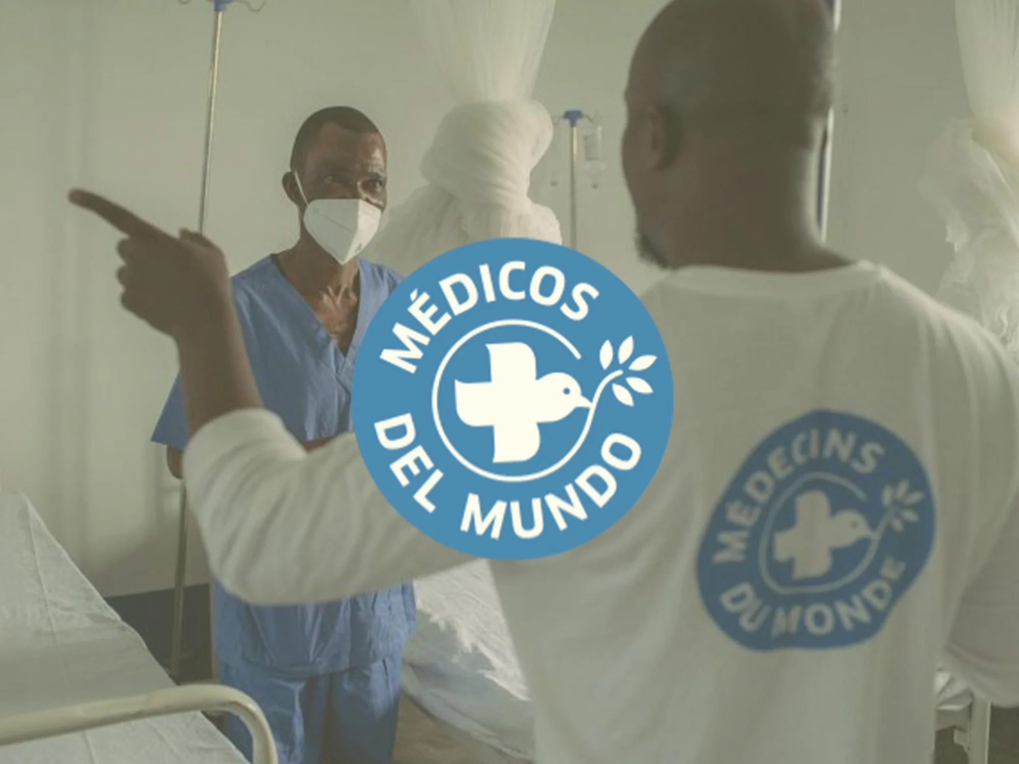 Un médico con mascarilla mira a través de un cristal, en un entorno hospitalario, con un símbolo de la Cruz y el texto 'MÉDICOS DEL MUNDO' en un círculo azul en el reflejo del cristal.