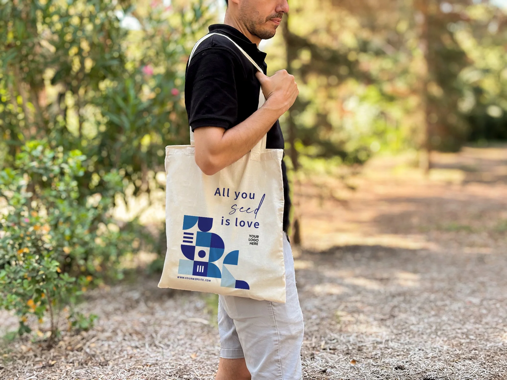Hombre en pantalones cortos usando una camiseta negra y cargando una bolsa de tela con texto y logotipo, en un sendero rodeado de naturaleza con árboles y follaje en el fondo.