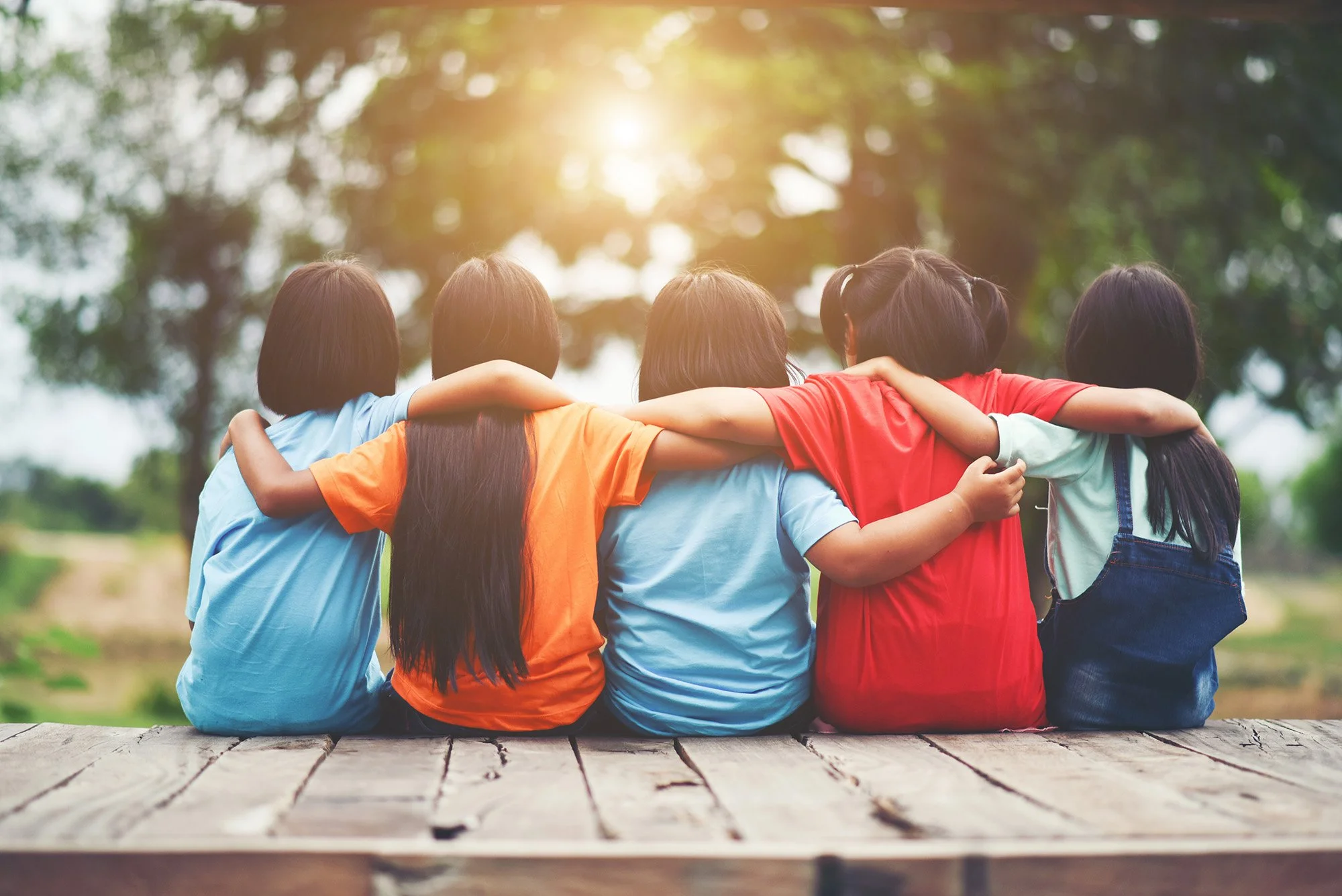 Un grupo de niños con camisetas de diferentes colores están abrazados, viendo hacia adelante en un parque con árboles, en un día soleado.