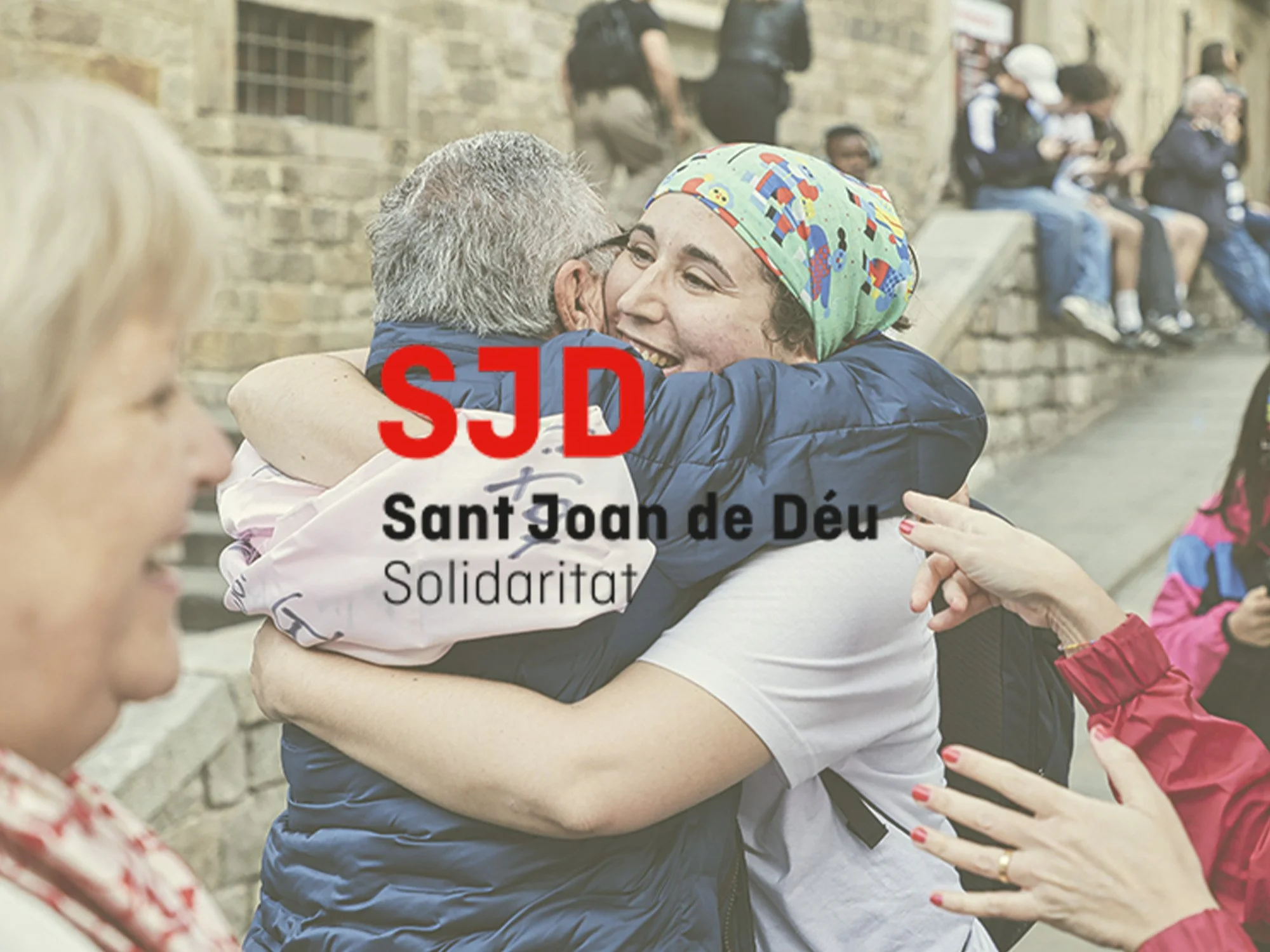 Dos personas abrazándose con alegría en una plaza, con varias personas sentadas en un muro de piedra en el fondo.