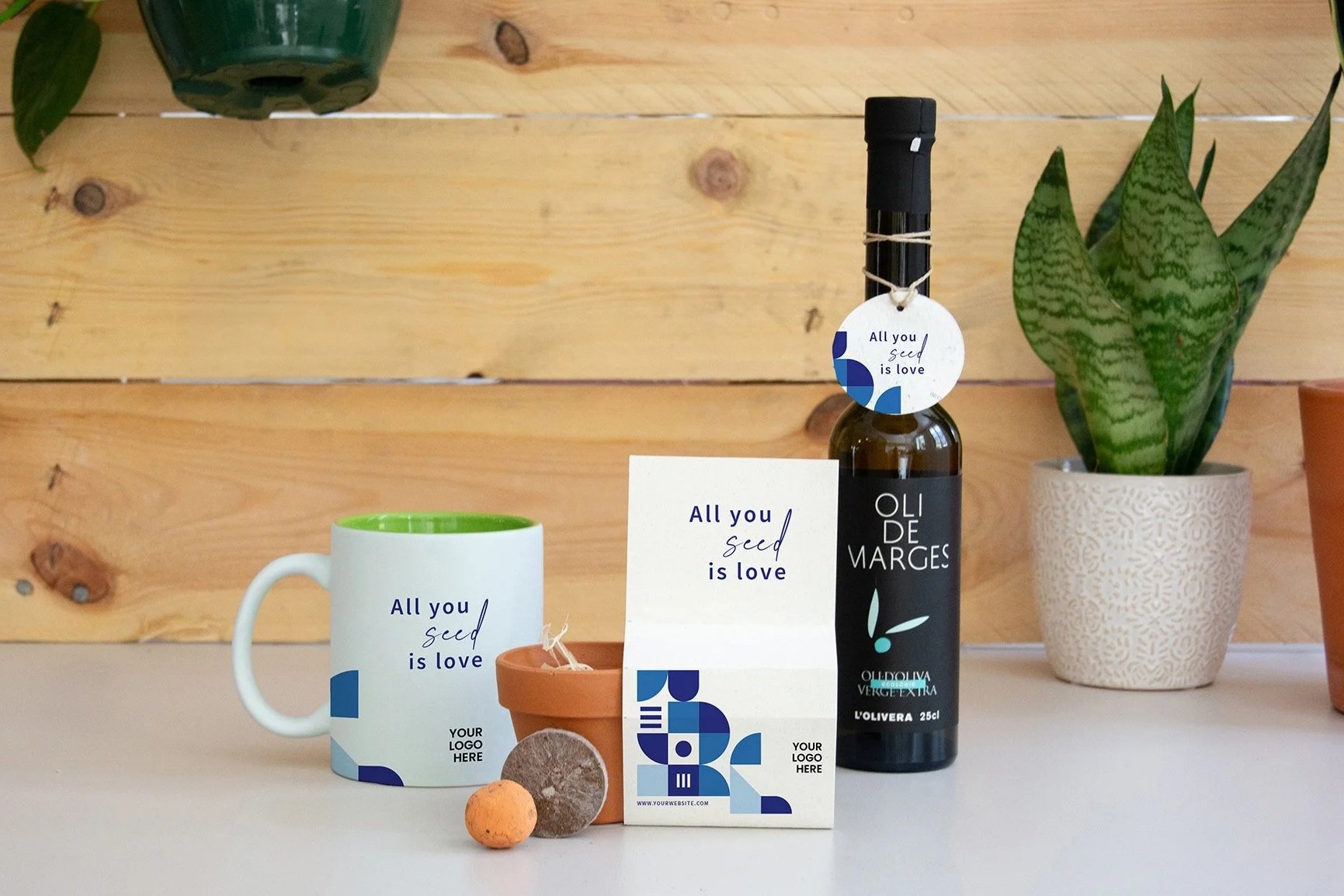 Botella de aceite de oliva con etiqueta que dice 'Oli de marges', taza blanca con interior verde y texto 'All you seed is love', tarjeta con el mismo mensaje, pequeño tiesto de barro con plantas verdes, y varias piedritas en una superficie de madera.