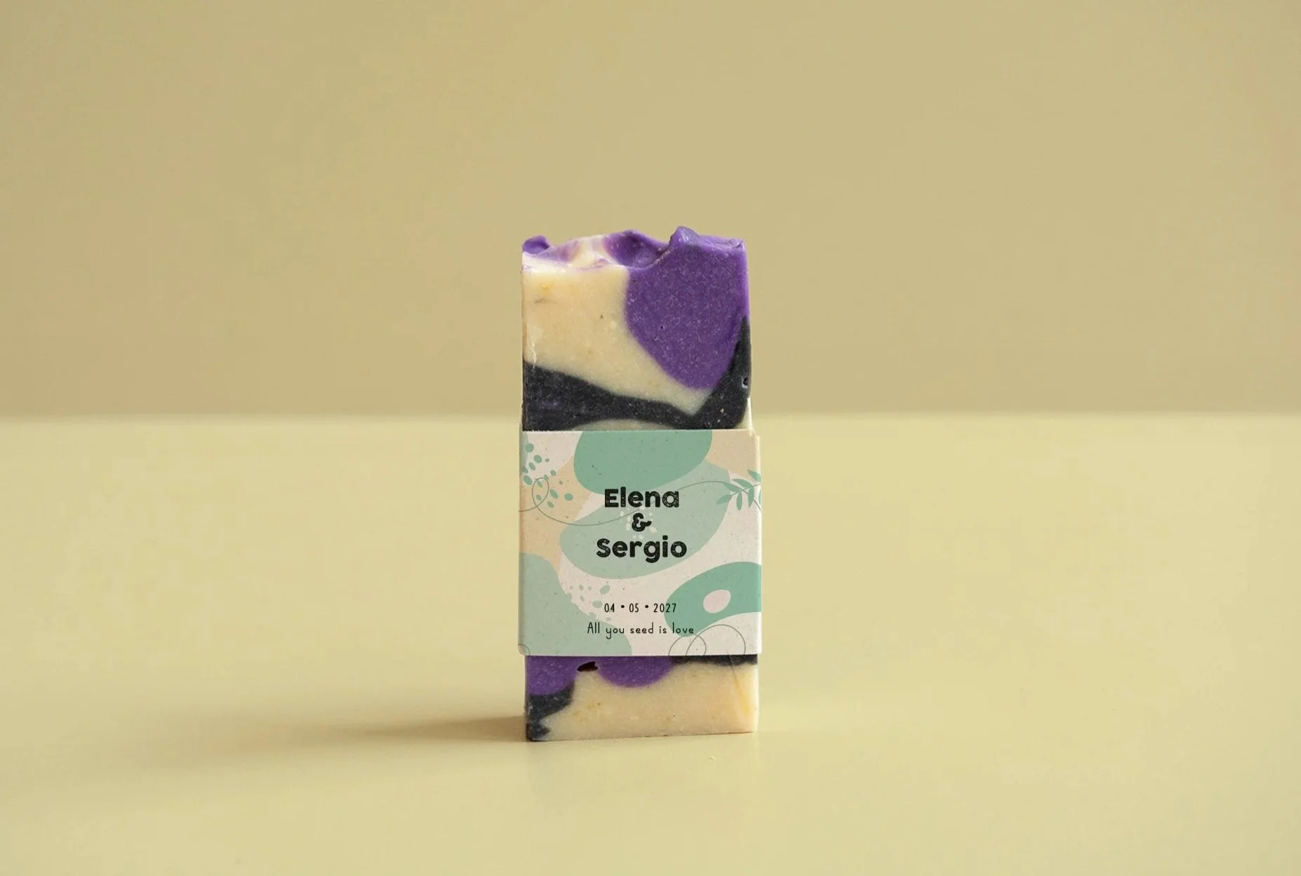 Jabón decorativo con diseño en tonos morados, blancos y negros, en una caja con etiquetas que dicen 'Elena & Sergio' y la fecha '04-05-2027', con la frase 'All you seed is love'