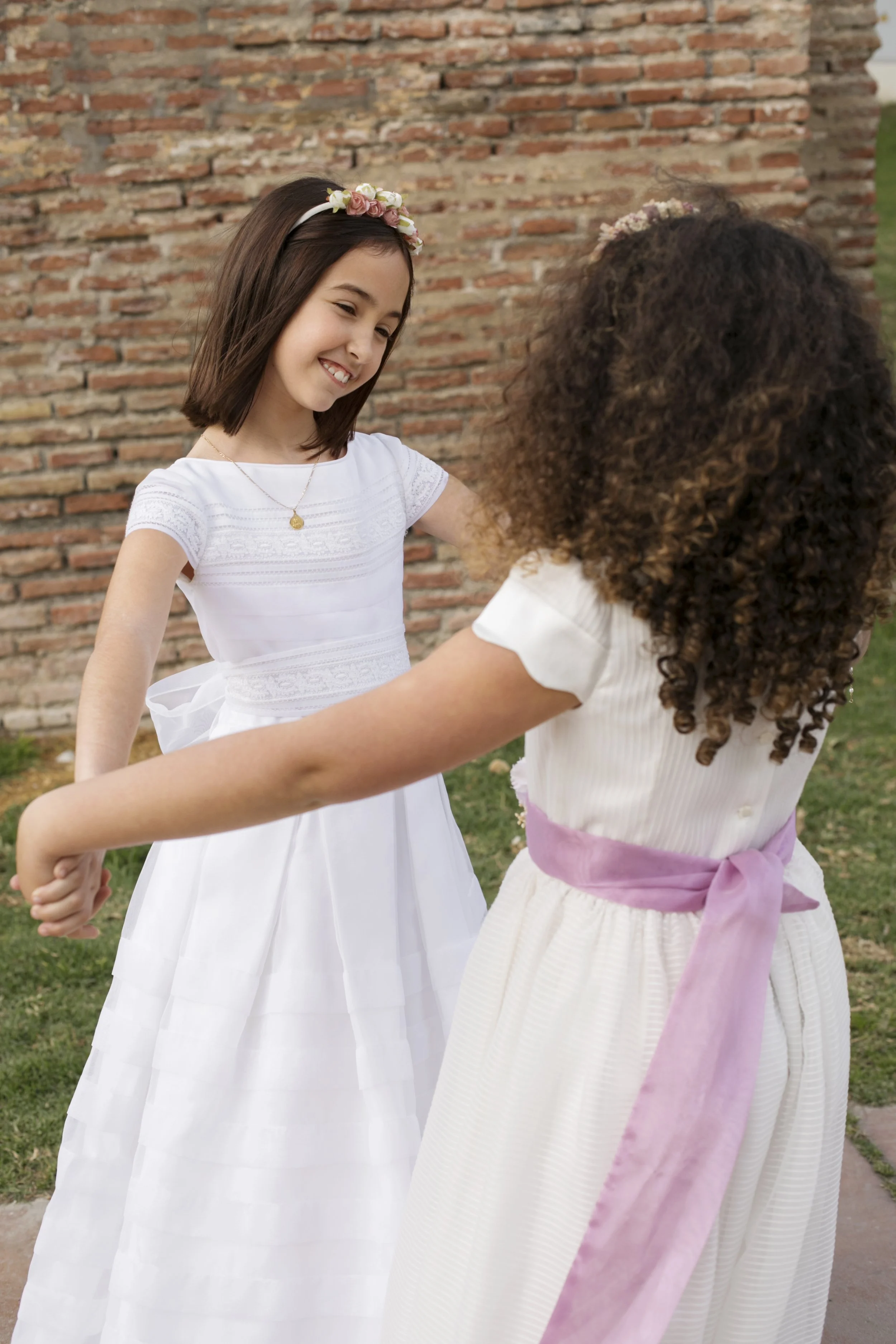 Dos niñas con vestidos blancos, una con una corona de flores en la cabeza y la otra con un lazo rosa en la cintura, parecen estar bailando o jugando juntas al aire libre con un muro de ladrillos y césped en el fondo.