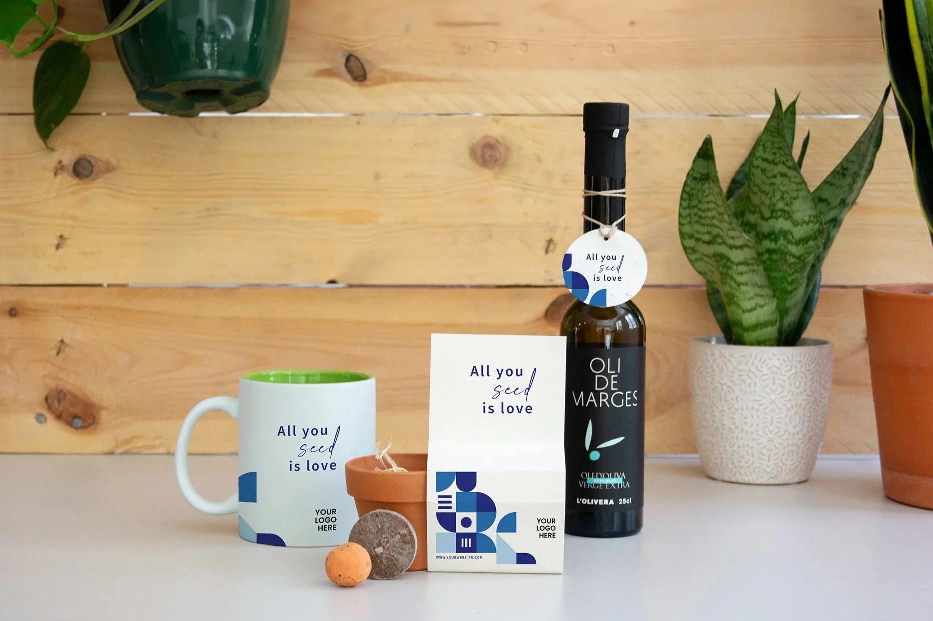 Varios objetos decorativos en una mesa de madera, incluyendo varias plantas en macetas, una botella de aceite de oliva, una taza con mensaje, y una tarjeta con texto 'All you seed is love'.