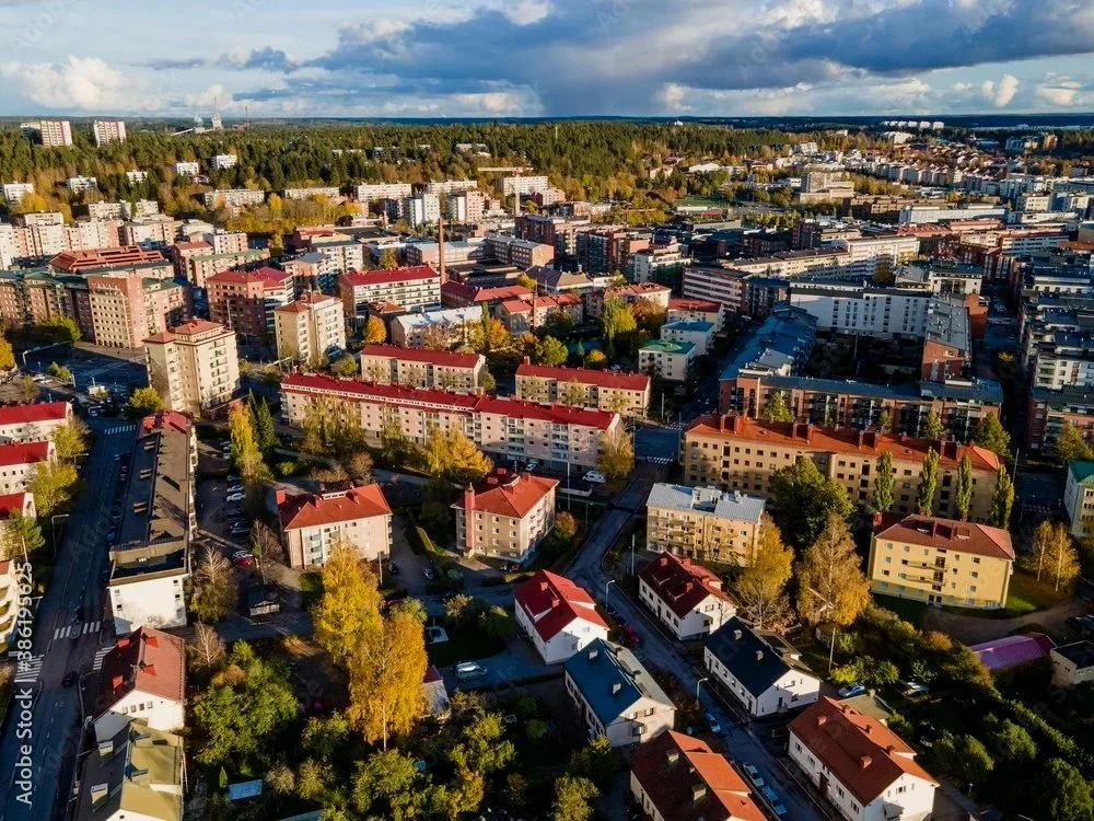Ilmasta otettu kuva kaupunkialueesta, jossa on useita rakennuksia, puita ja teitä, sekä taivaalla pilviä.