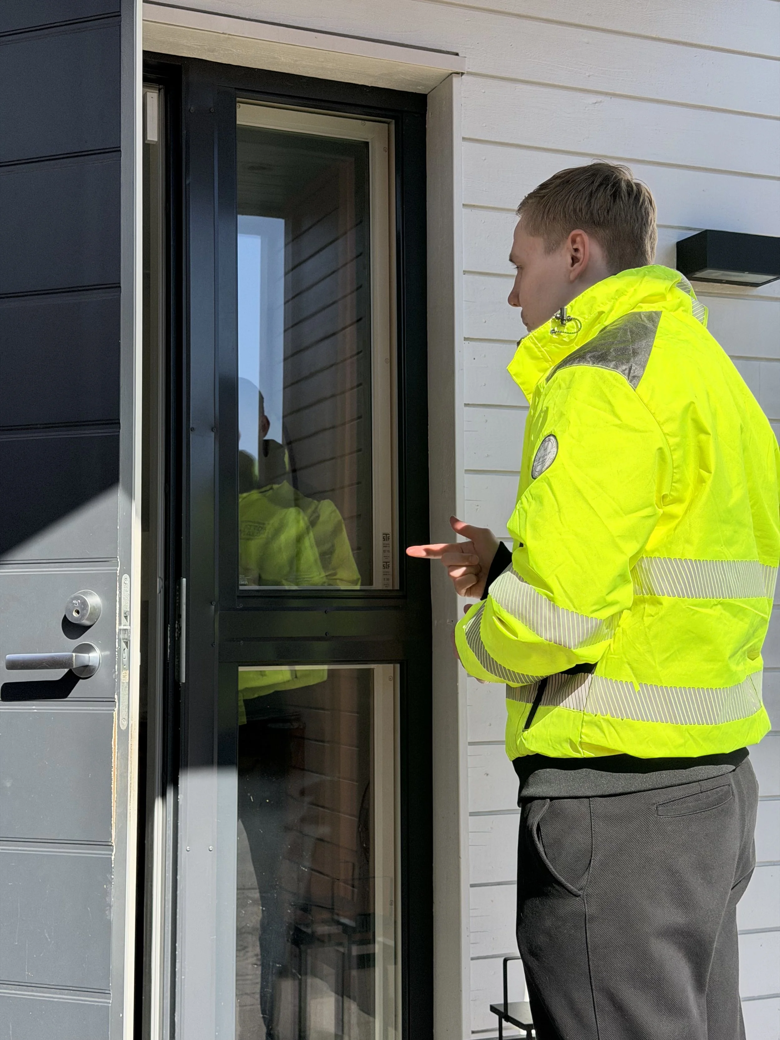 Mies pukeutuneena kirkkaankeltaiseen takkiin ja mustiin housuihin seisoo ulkona kädet ristissä, katsellen asuntoa ulko-oven suuntaan.