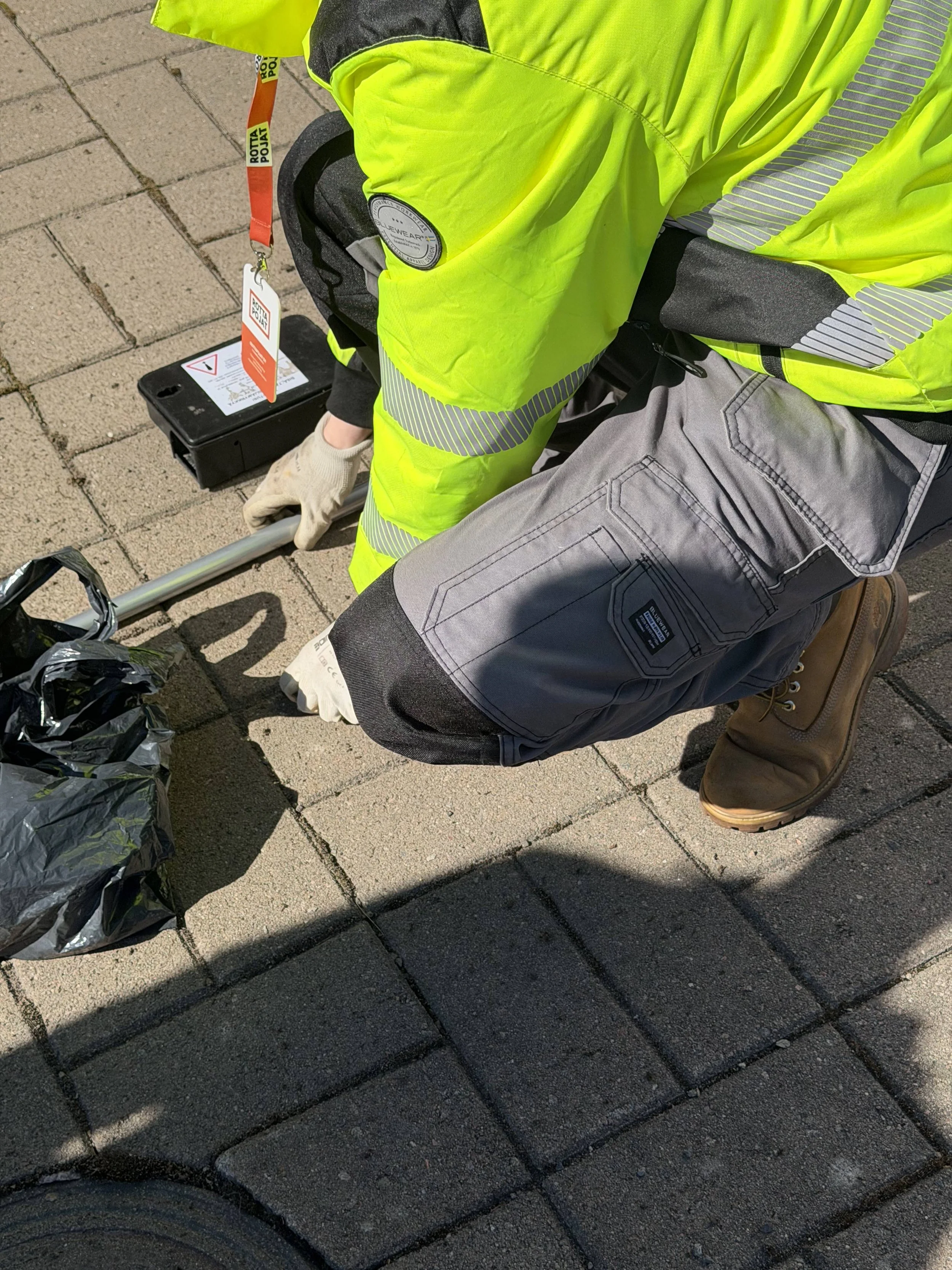 Kuvassa näkyy henkilö, joka pukeutuneena keltaiseen huomioliiviin ja harmaihin työvaatteisiin kerää roskia katujalasta, jossa on jonkin verran roskia, kuten mustia muovipusseja.