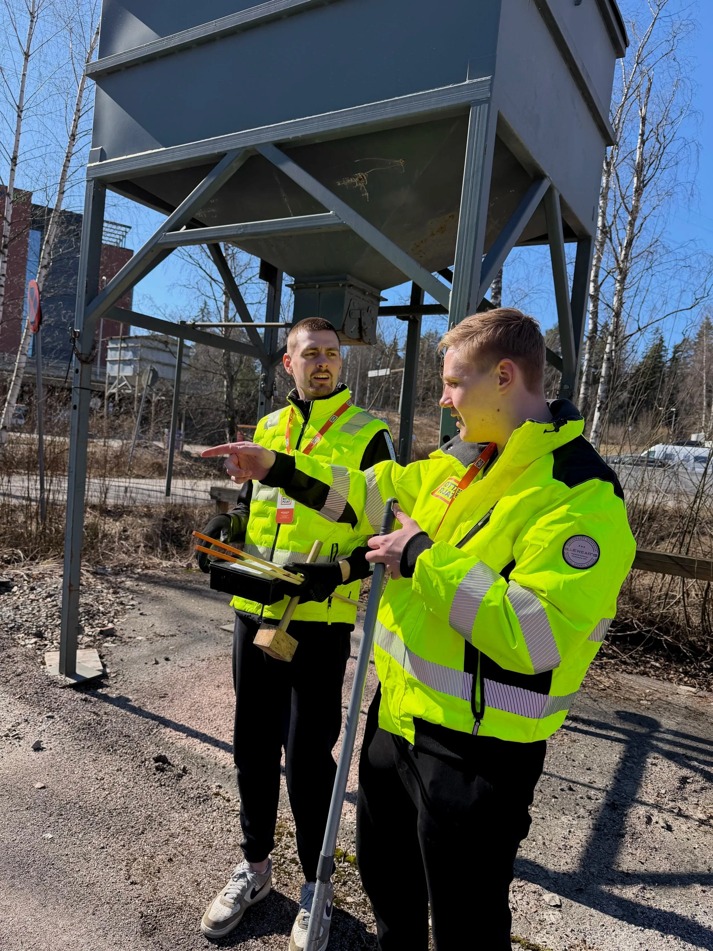 Kaksi miestä keskustelee ulkona, molemmilla yllään keltaiset huomioväriset takit ja yläosat, toinen mies osoittaa sormellaan ja pitää kansiota ja ulkoiluvälineitä, taustalla rakennus ja puut.