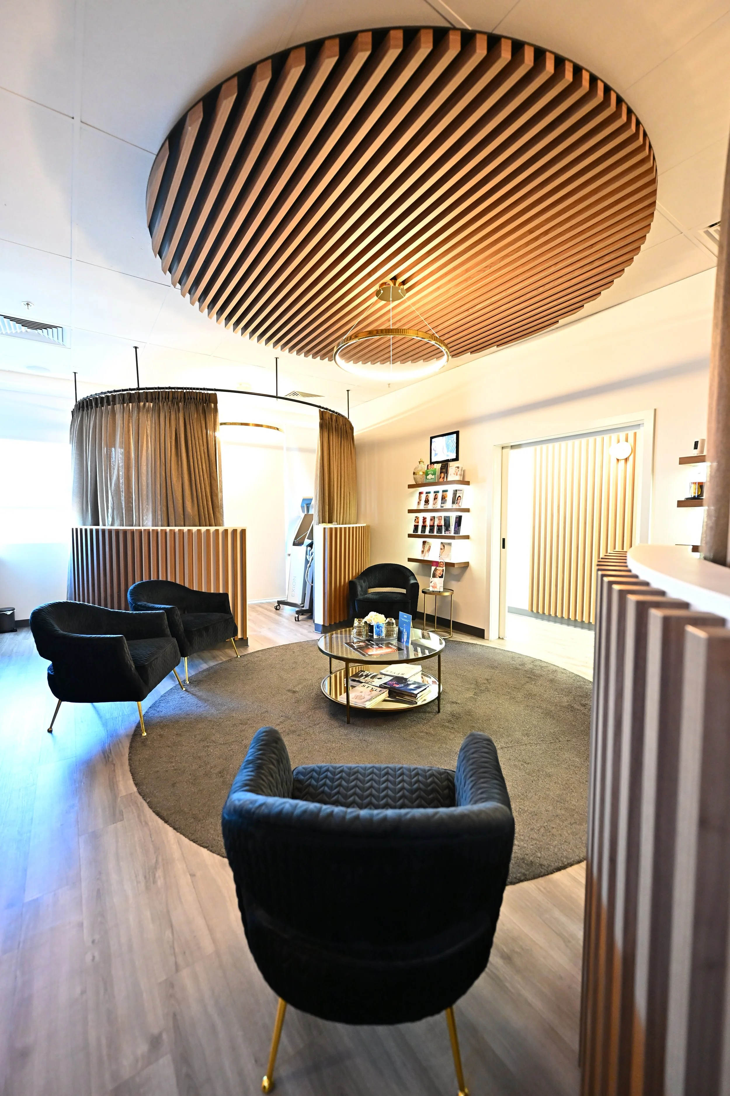 Modern office or waiting room with black armchairs, a round coffee table with magazines, a circular wooden ceiling feature, and beige curtains creating a semi-private space.