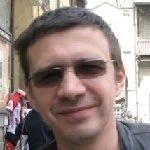 A man with short dark hair wearing sunglasses, smiling outdoors in front of a building.