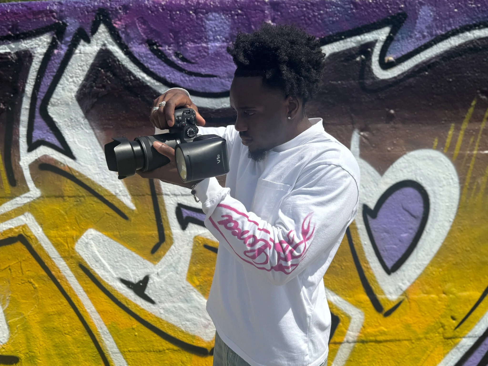 A person with curly black hair taking photos with a camera in front of a colorful graffiti wall with purple, yellow, white, and black abstract shapes and hearts.