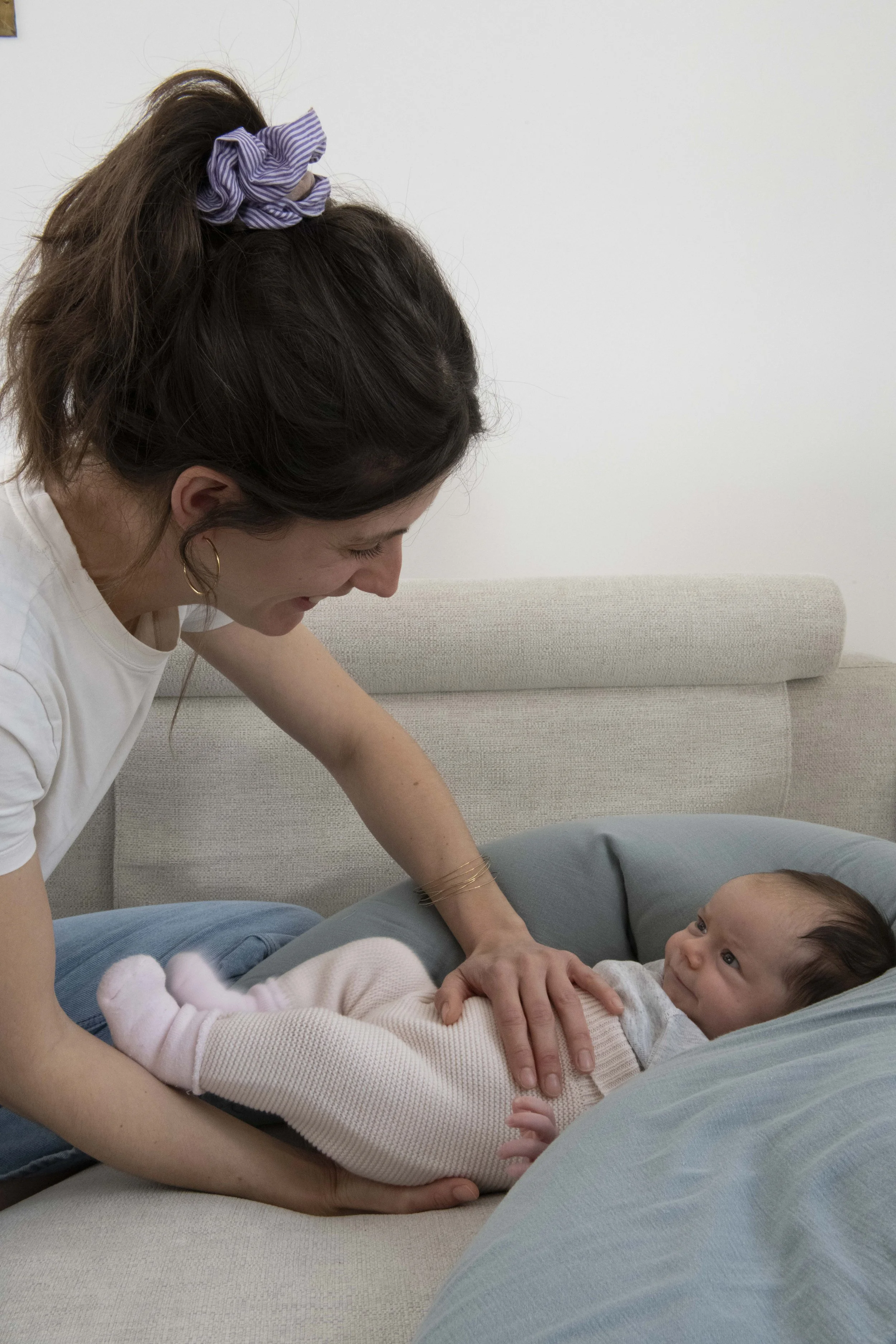 Une femme souriante joue avec un bébé allongé sur un canapé, dans une pièce aux murs blancs.