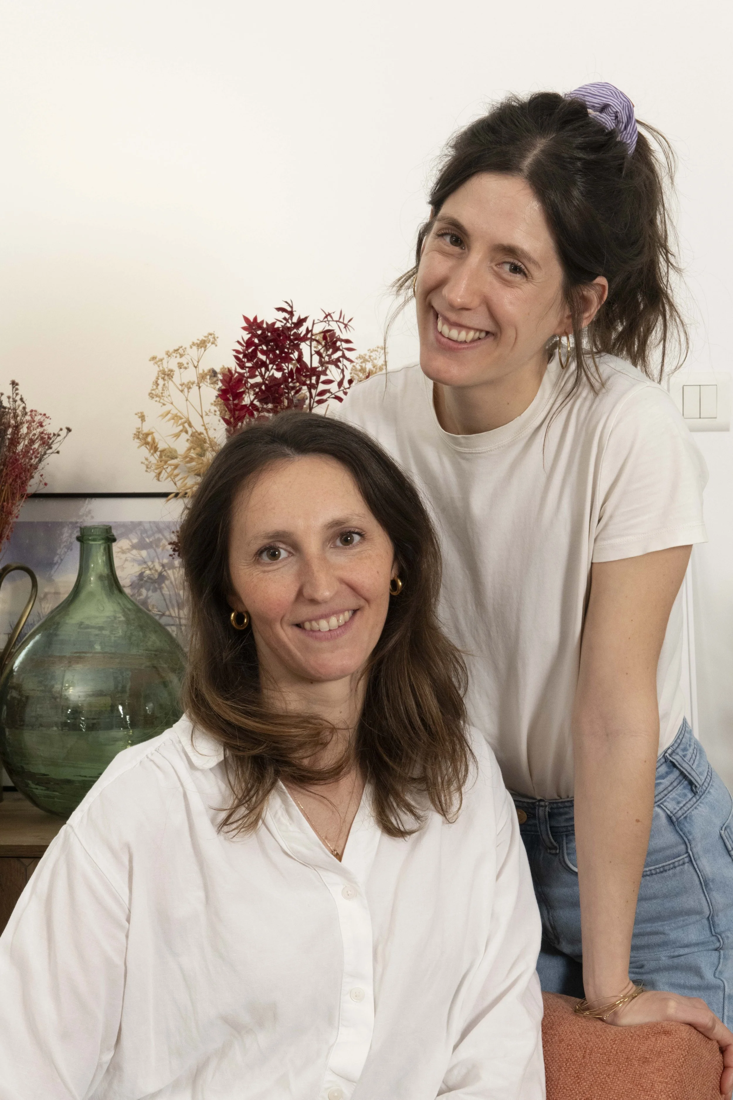 Deux femmes souriantes posant dans un cadre intérieur, avec des fleurs séchées en arrière-plan et une grande jarre en verre à côté d'elles.