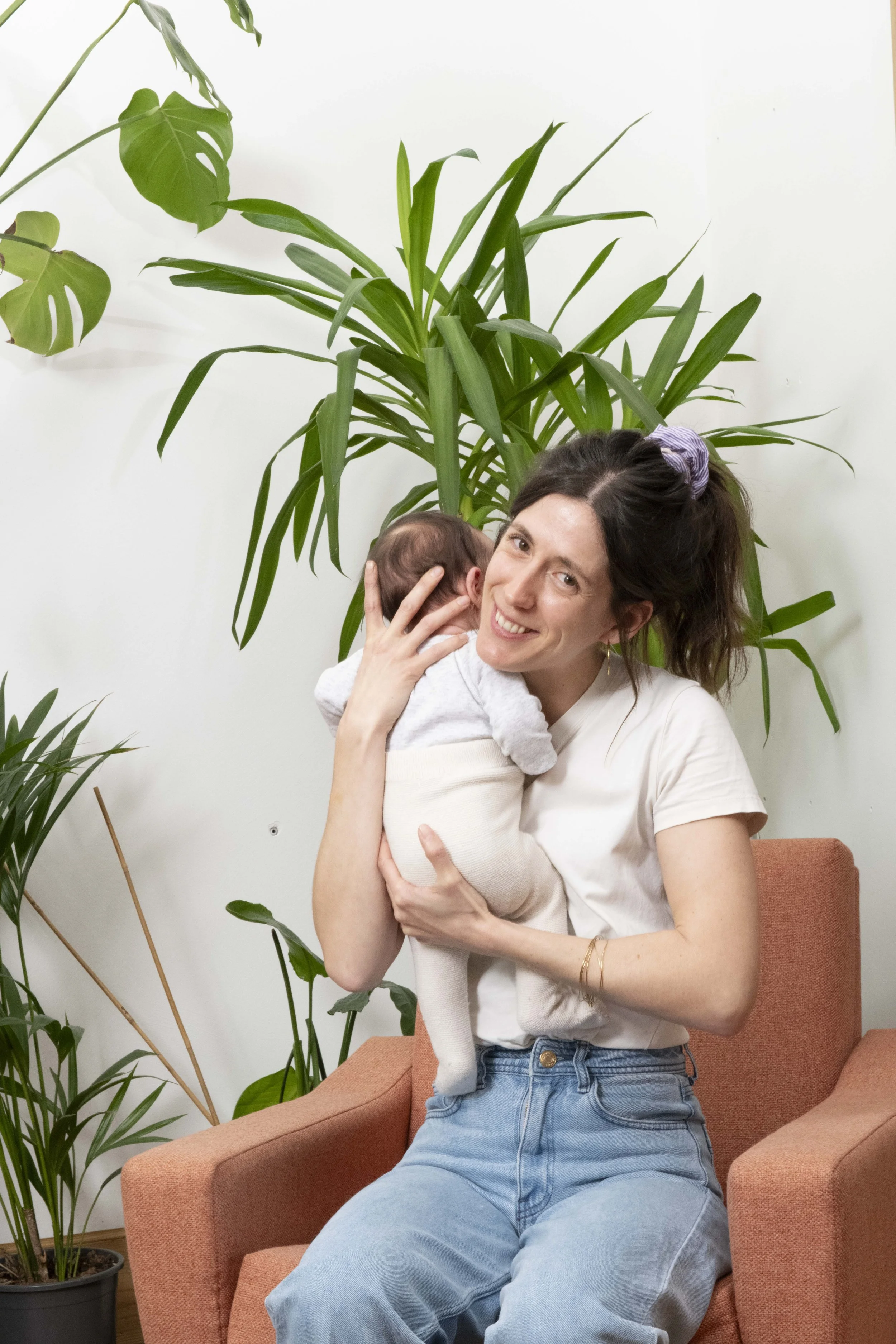 Une femme souriante tient un bébé dans ses bras, assise sur un fauteuil orange en présence de plusieurs plantes vertes contre un mur blanc.