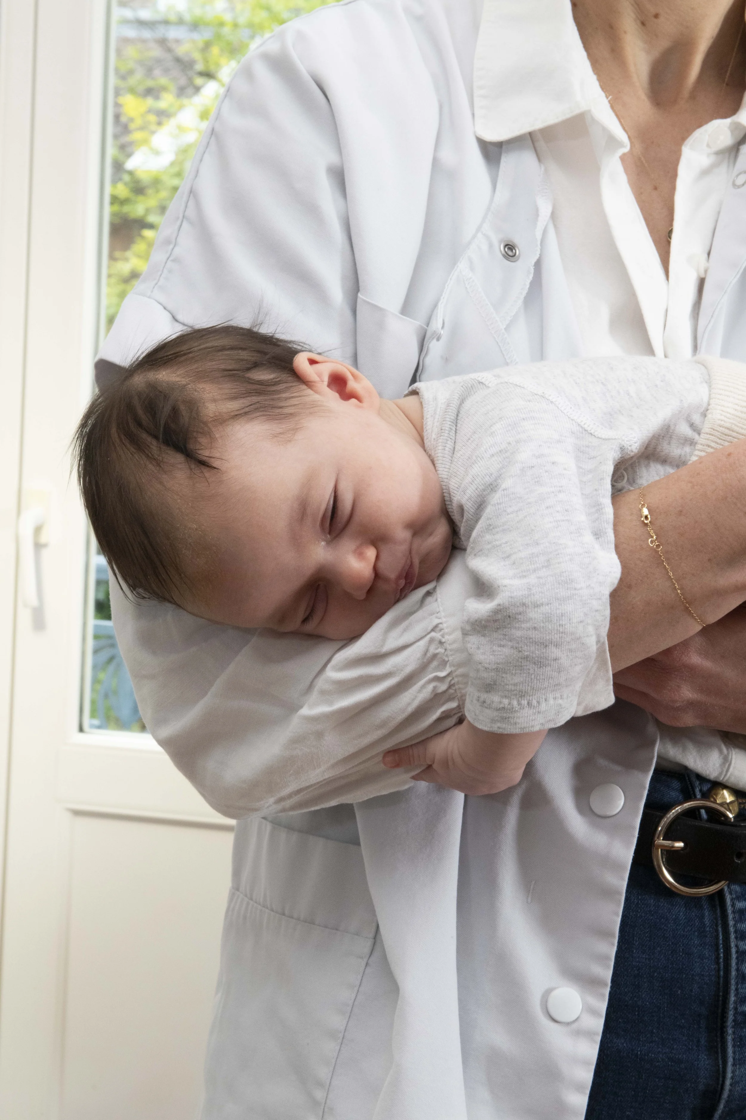 Un enfant est allongé sur une table ou un lit, en train de dormir, sur une personne en blouse blanche, probablement un professionnel de santé, dans une pièce lumineuse avec une fenêtre en arrière-plan.