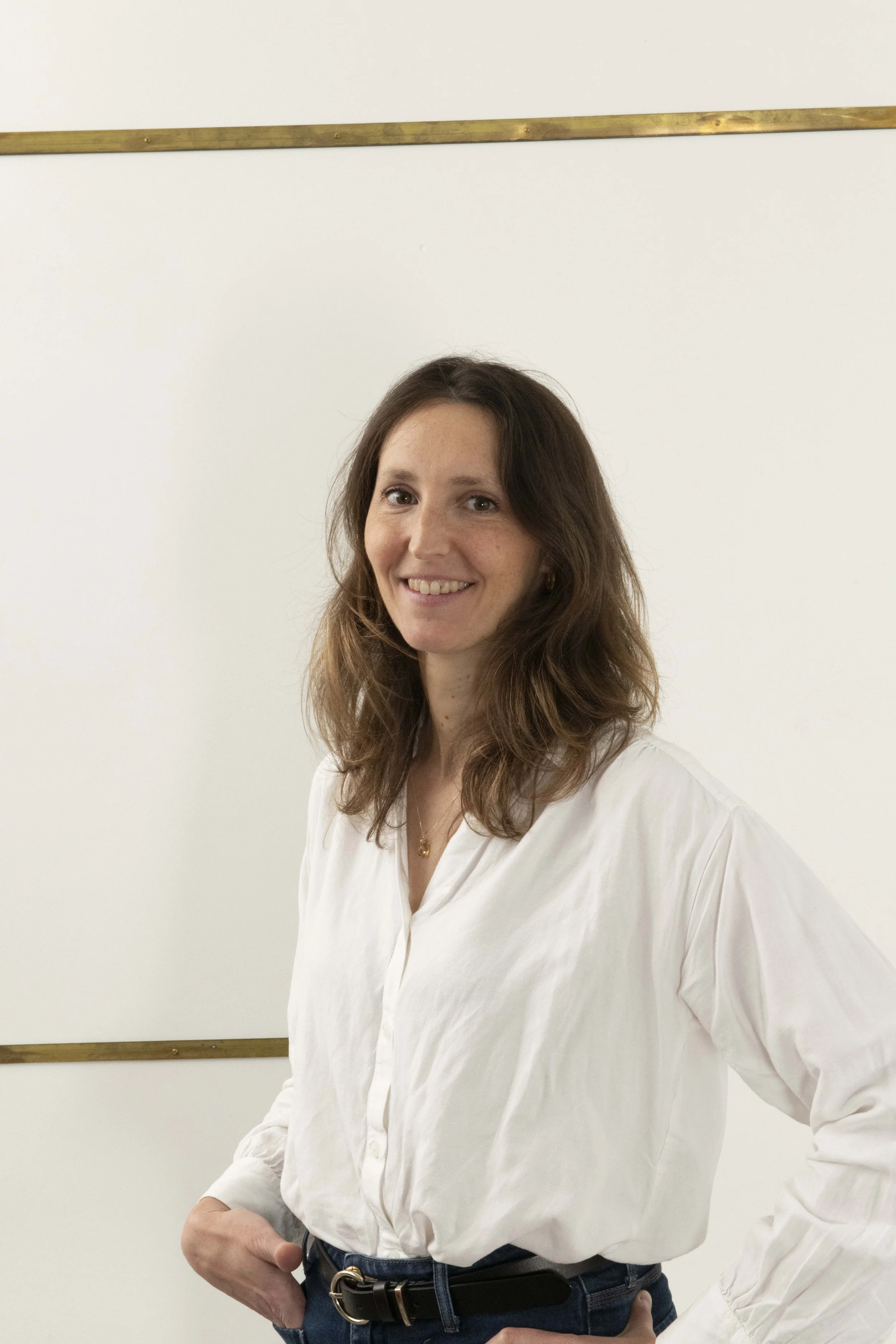 Une femme avec des cheveux bruns, portant une chemise blanche à manches longues, sourit devant un tableau blanc dans une salle
