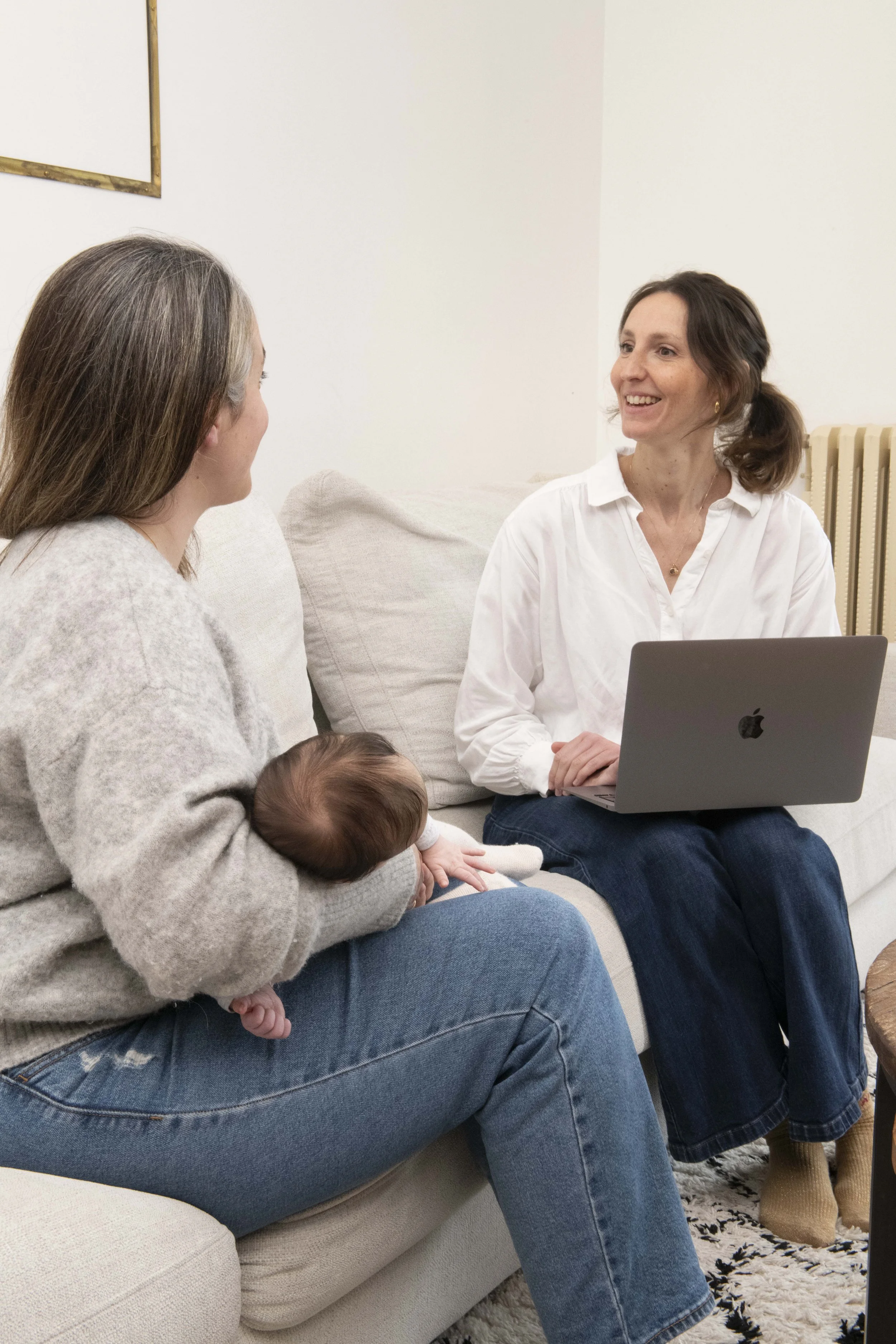 Deux femmes discutent sur un canapé en présence d'un bébé, avec un ordinateur portable sur les genoux d'une d'elles, dans une pièce au décor simple.