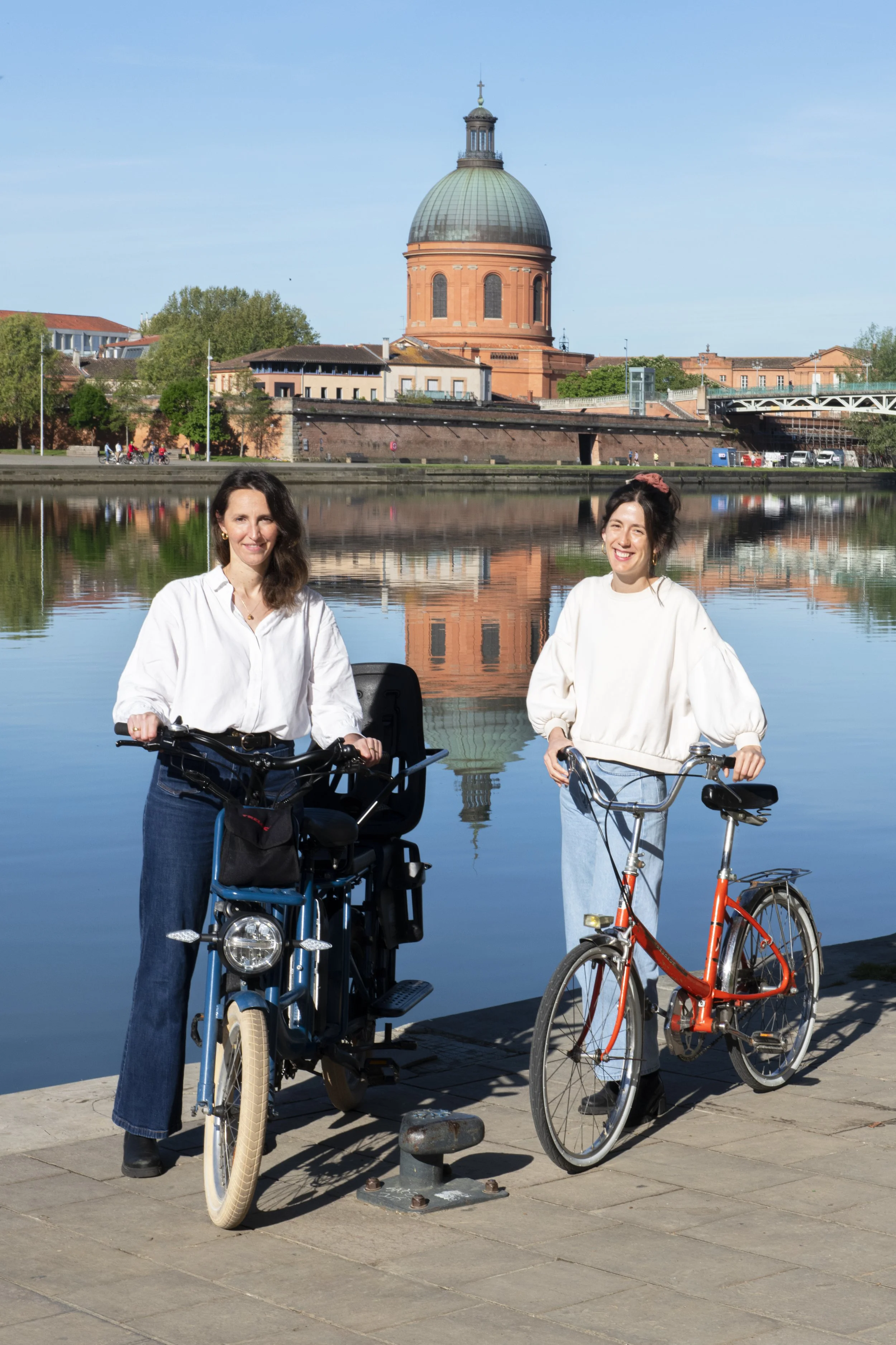 Deux femmes posent avec leurs vélos au bord d'une rivière, avec un bâtiment historique à dôme en arrière-plan sous un ciel clair.