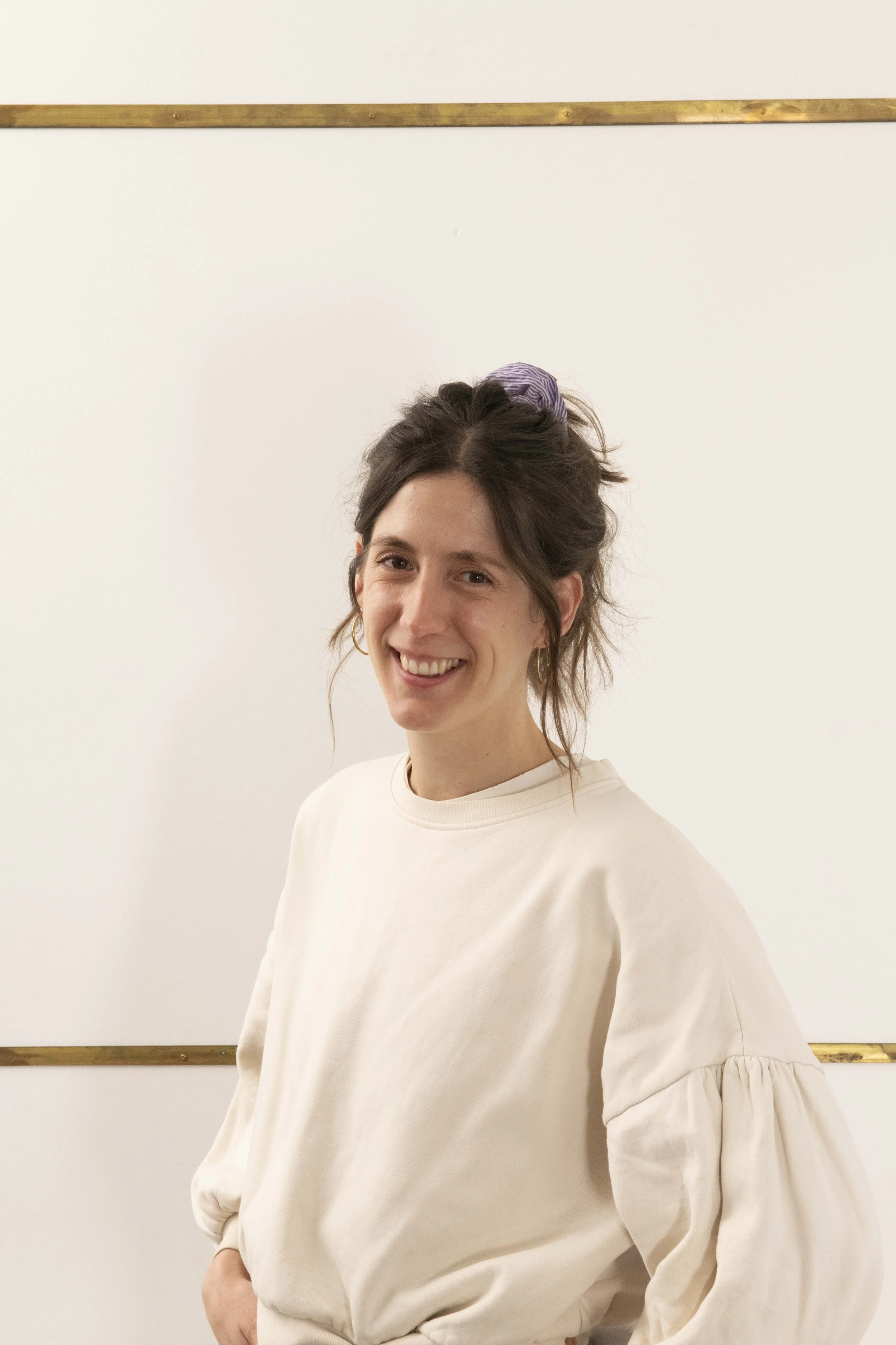 Jeune femme souriante avec des cheveux bruns attachés avec un foulard violet, portant un sweat blanc, se tenant devant un tableau blanc dans une salle