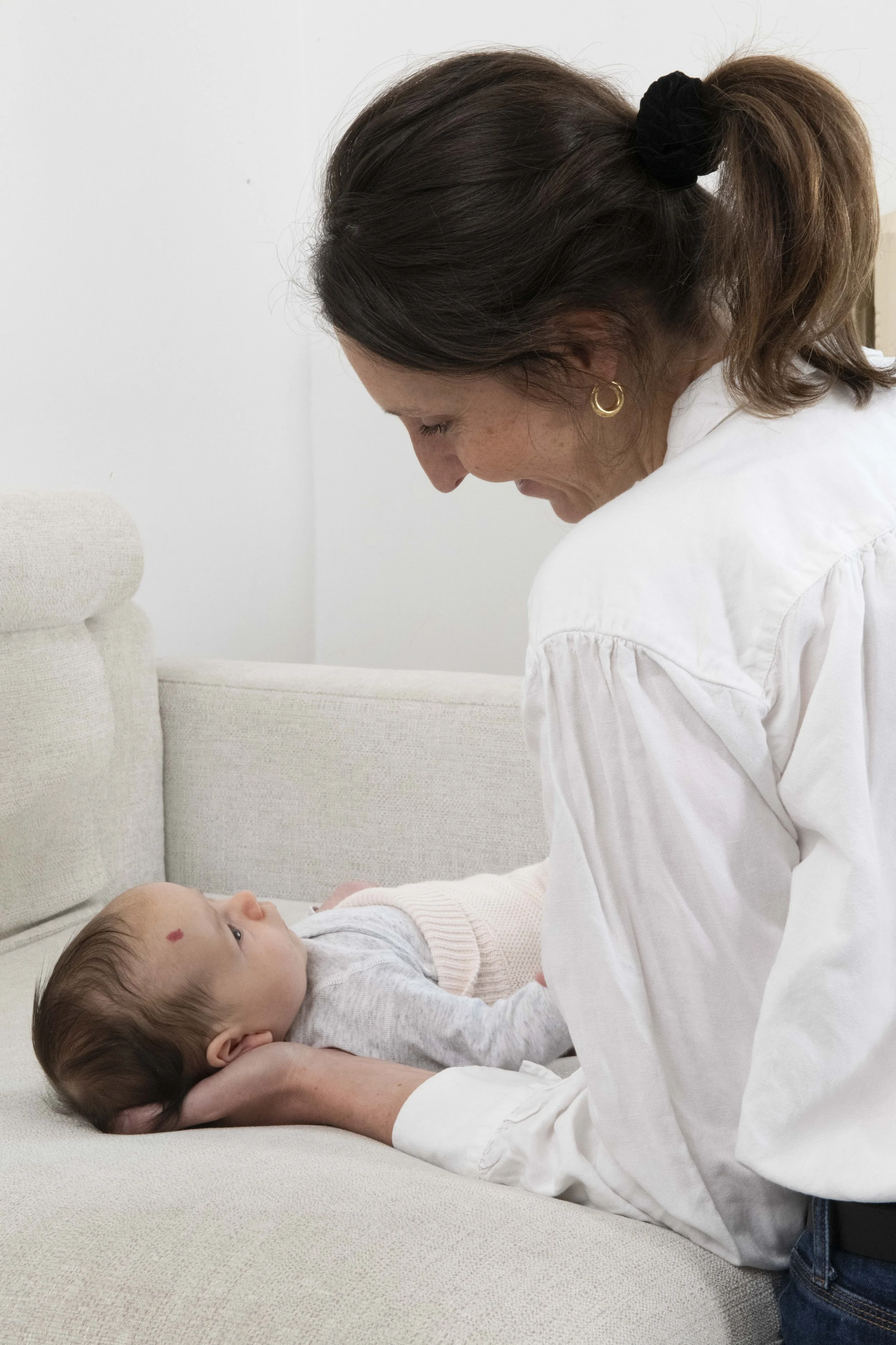Une femme avec des boucles d'oreilles dorées tient un bébé allongé sur un canapé beige. La femme porte une chemise blanche et regarde tendrement l'enfant, qui regarde vers la caméra avec une tache rouge sur le front.