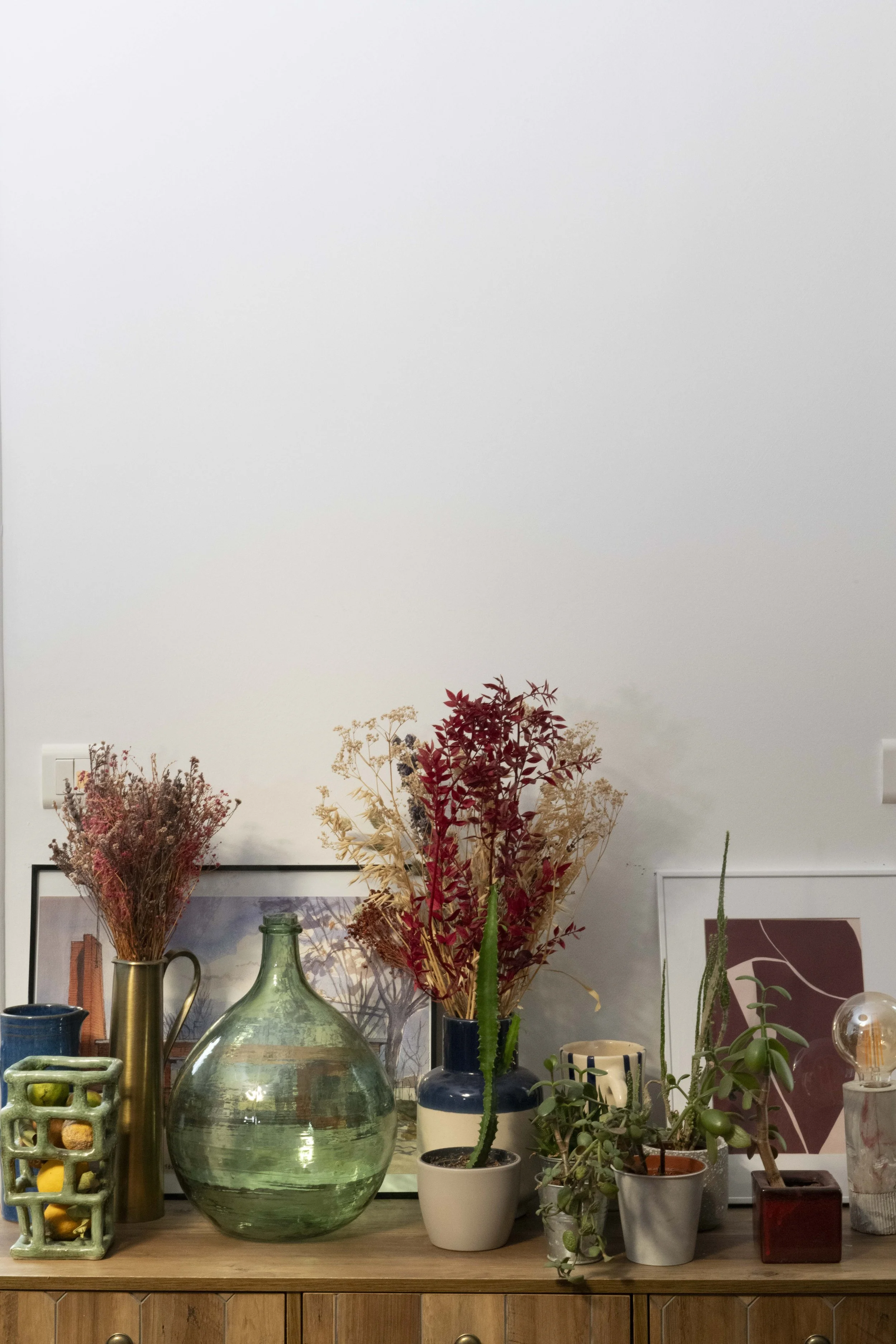 Vase en verre transparent avec une plante cactus, un bouquet de fleurs séchées dans un récipient bleu, plusieurs autres plantes en pots, un tableau et des objets décoratifs sur une table en bois contre un mur blanc.