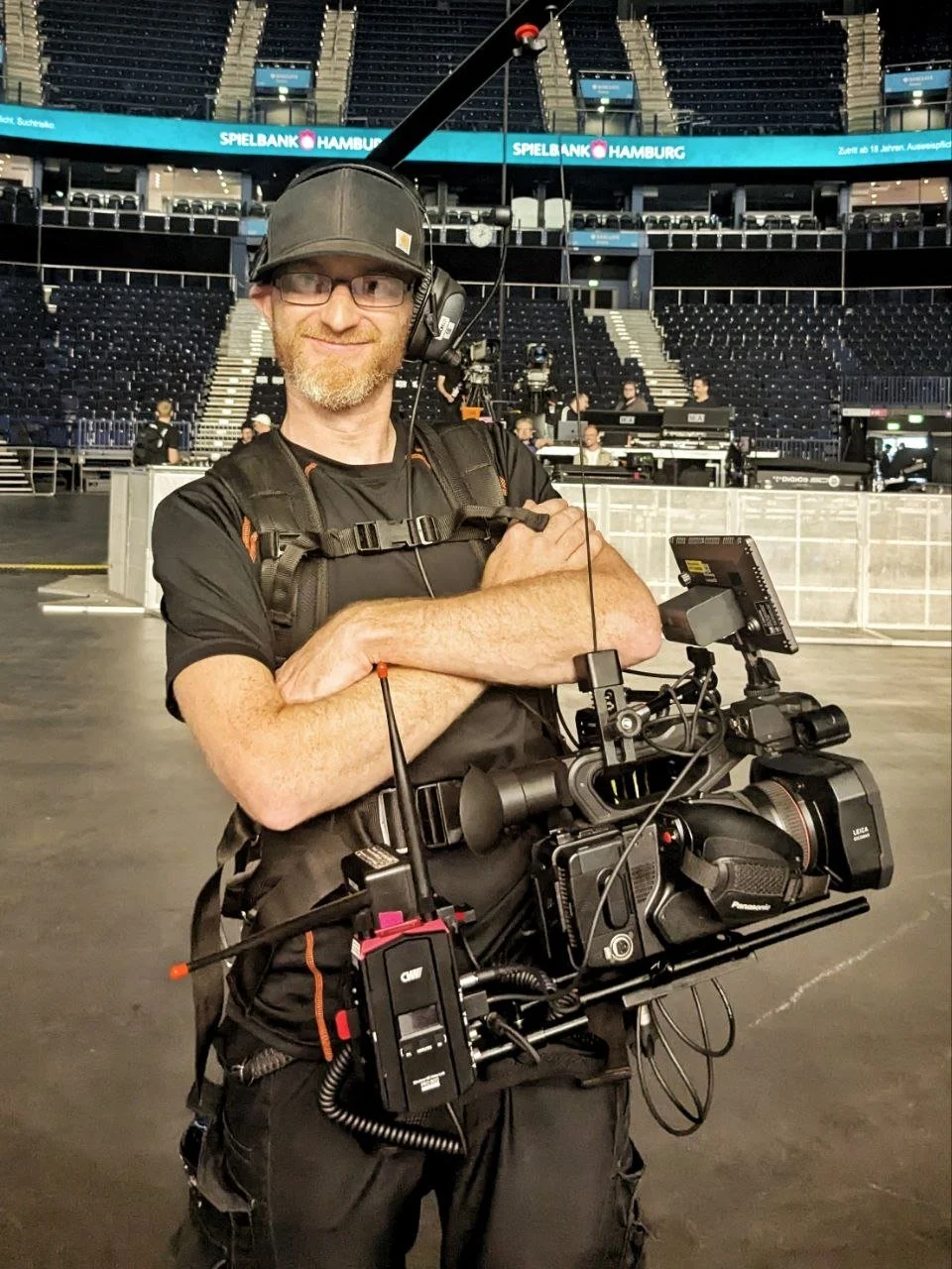 A man wearing glasses and a baseball cap standing in an arena with a professional video camera rig, with empty seats and some people working in the background.