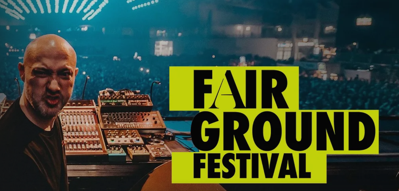 A DJ with a shaved head and beard making a face, standing behind DJ equipment at a music festival, with a large crowd and stage lights in the background, advertising the Fair Ground Festival.