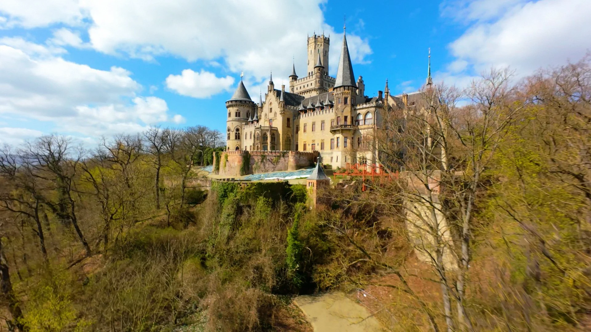 A large fairy-tale castle on a hill surrounded by trees and a cloudy blue sky.