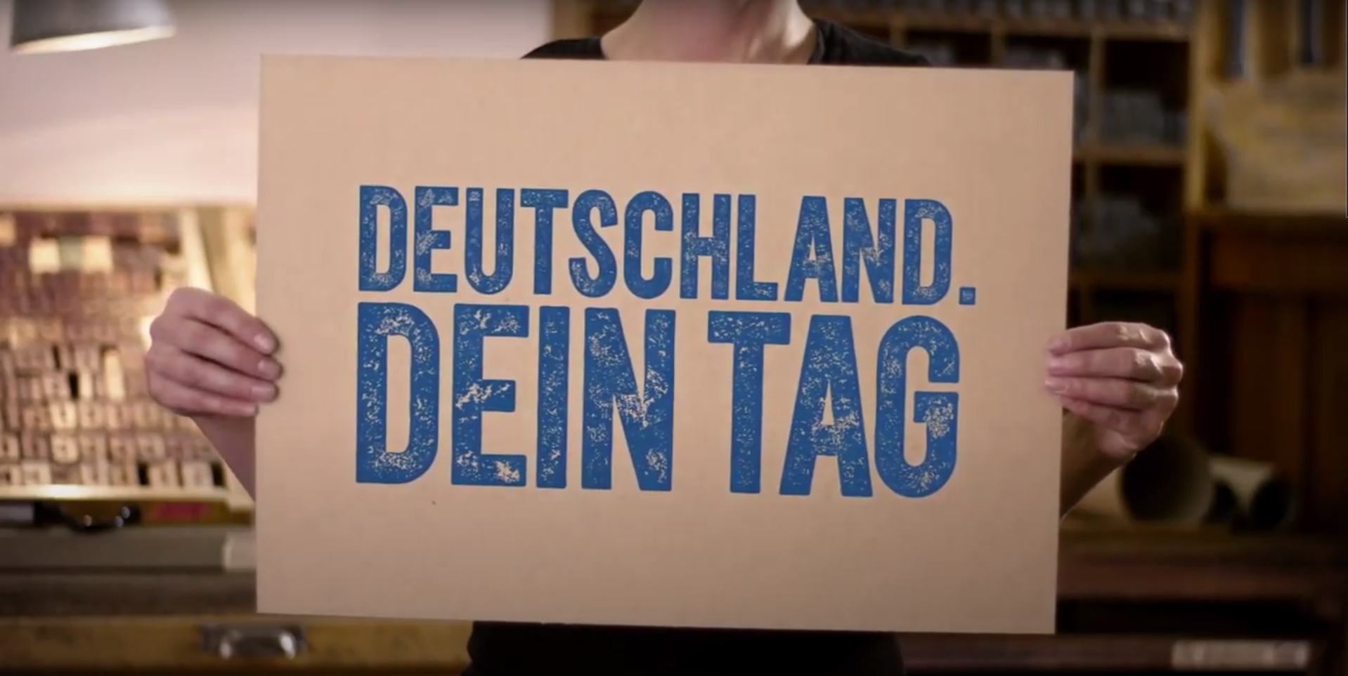 Person holding a beige sign with blue text that reads "Deutschland. Dein Tag" in a room with wooden shelves and storage.