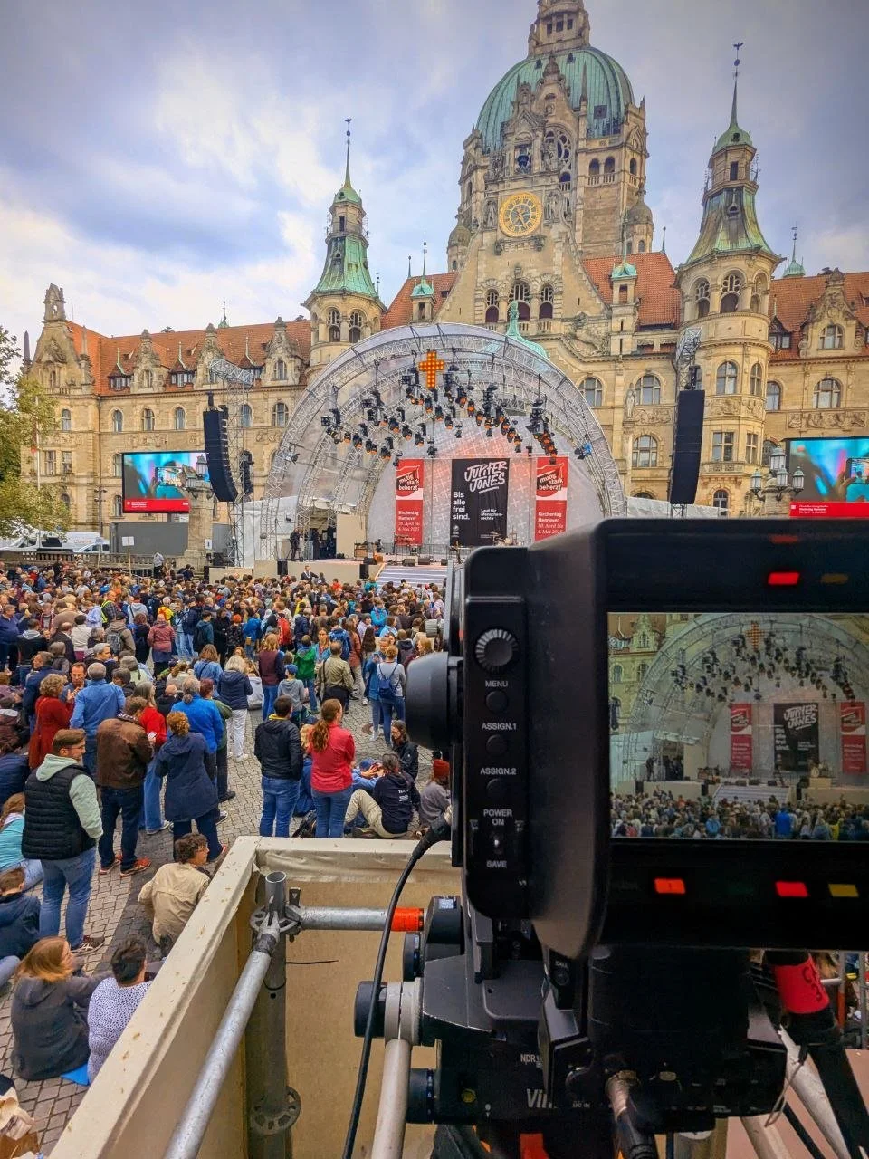 Outdoor concert at a historic building with a large crowd watching a stage. The stage has a canopy and screens with banners that say "JUPITER JONES" and advertisements.
