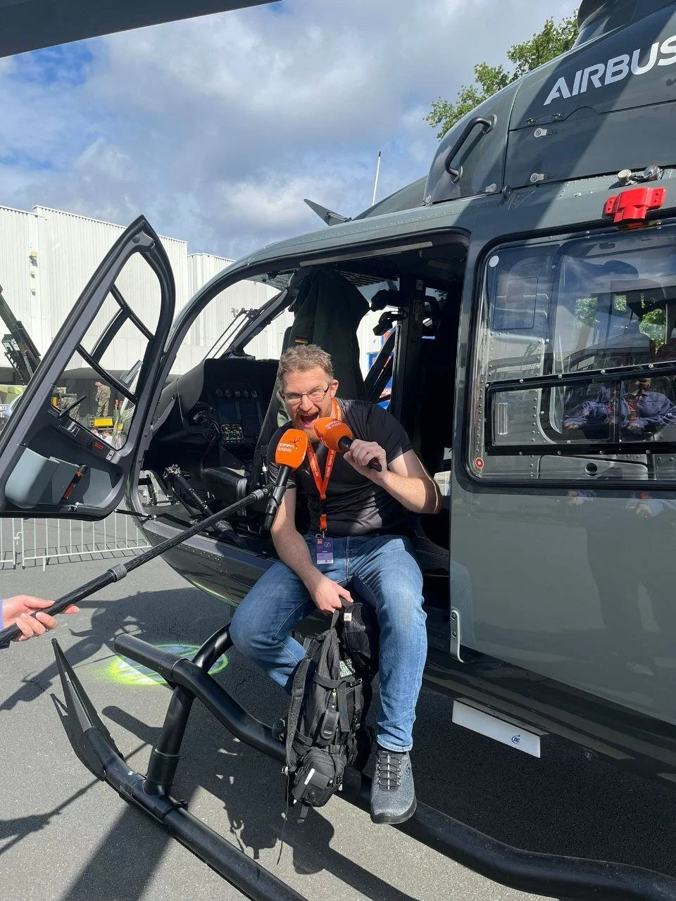A man sitting in the open door of a helicopter, holding two orange microphones, speaking animatedly, with a white building and partly cloudy sky in the background.