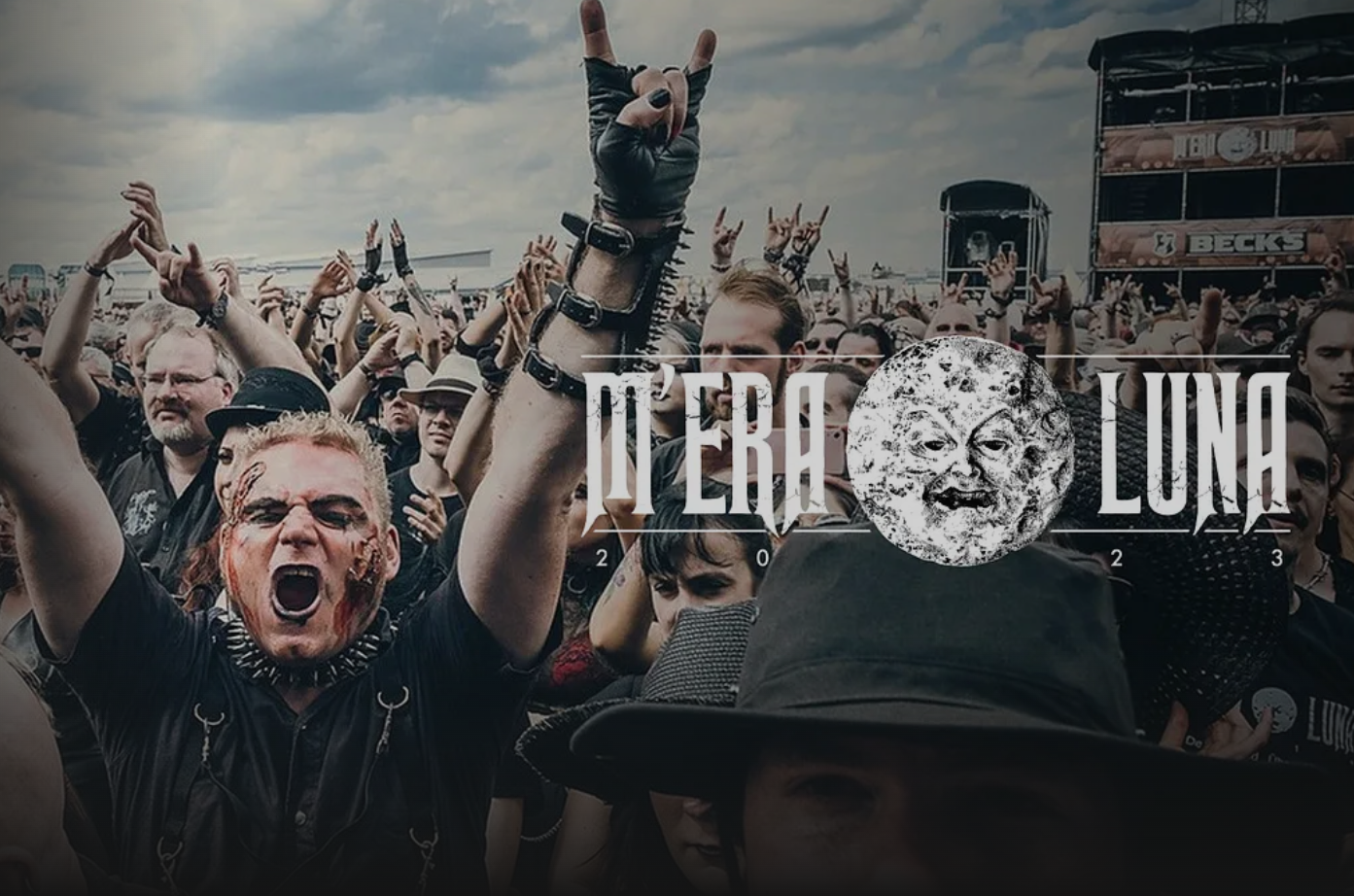 Crowd at a rock concert with many people raising their hands and shouting, some wearing dark clothing, while a person in the foreground has face makeup and an aggressive expression, with a large stage and speaker systems in the background.